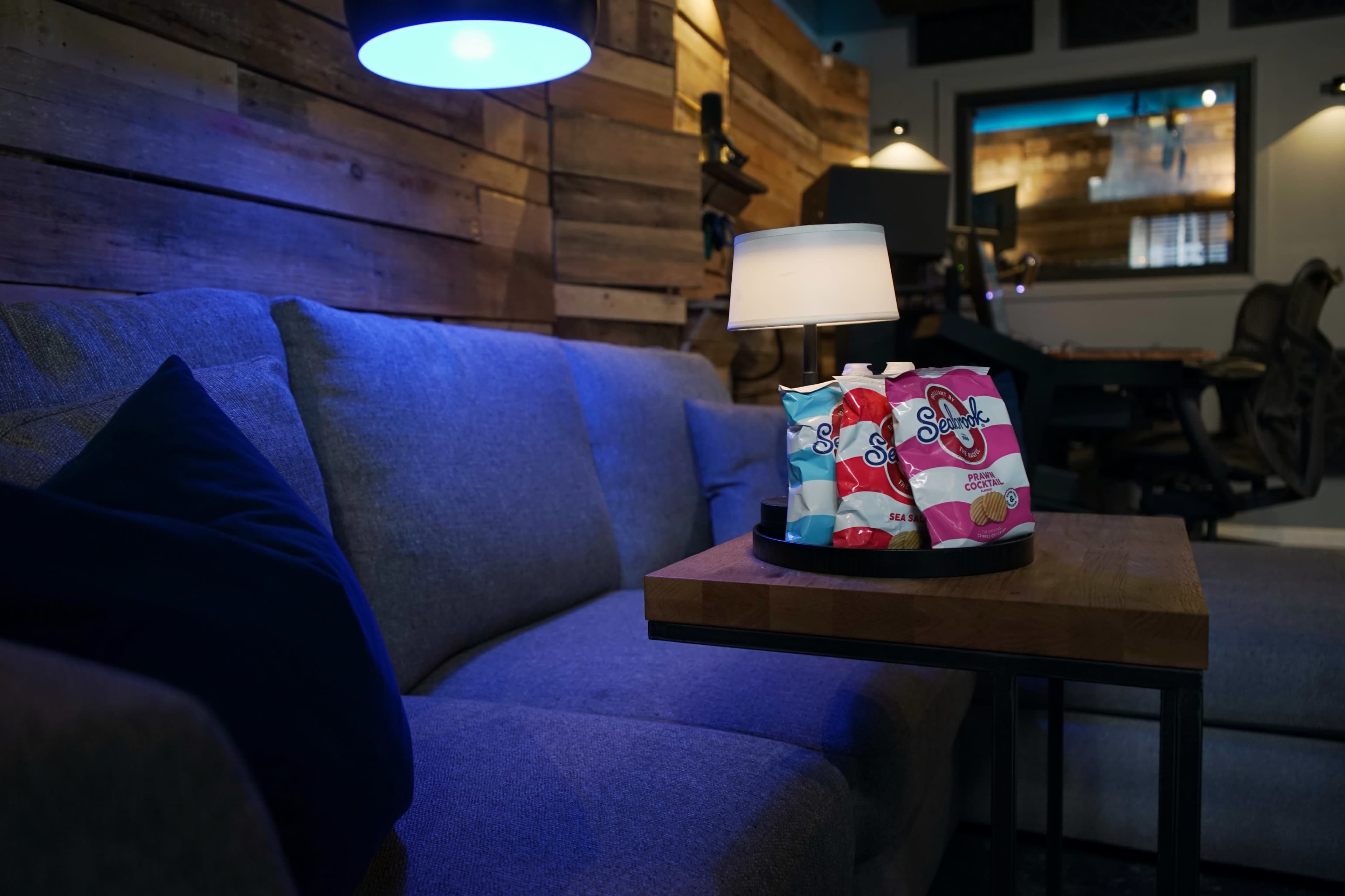 A gray sofa is positioned next to a wooden table holding colorful snack bags under a lamp in a cozy room with wooden walls.