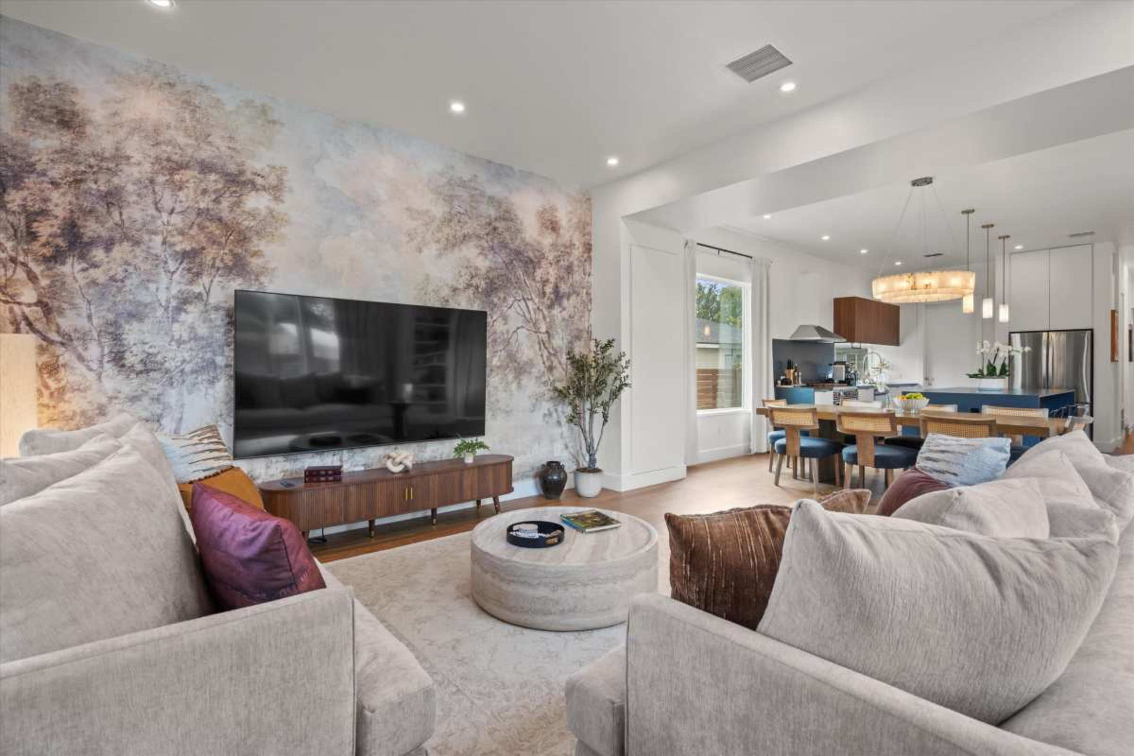 The image shows a modern living room with a large TV, plush sofas, and a round coffee table, connected to a dining area with a kitchen in the background.