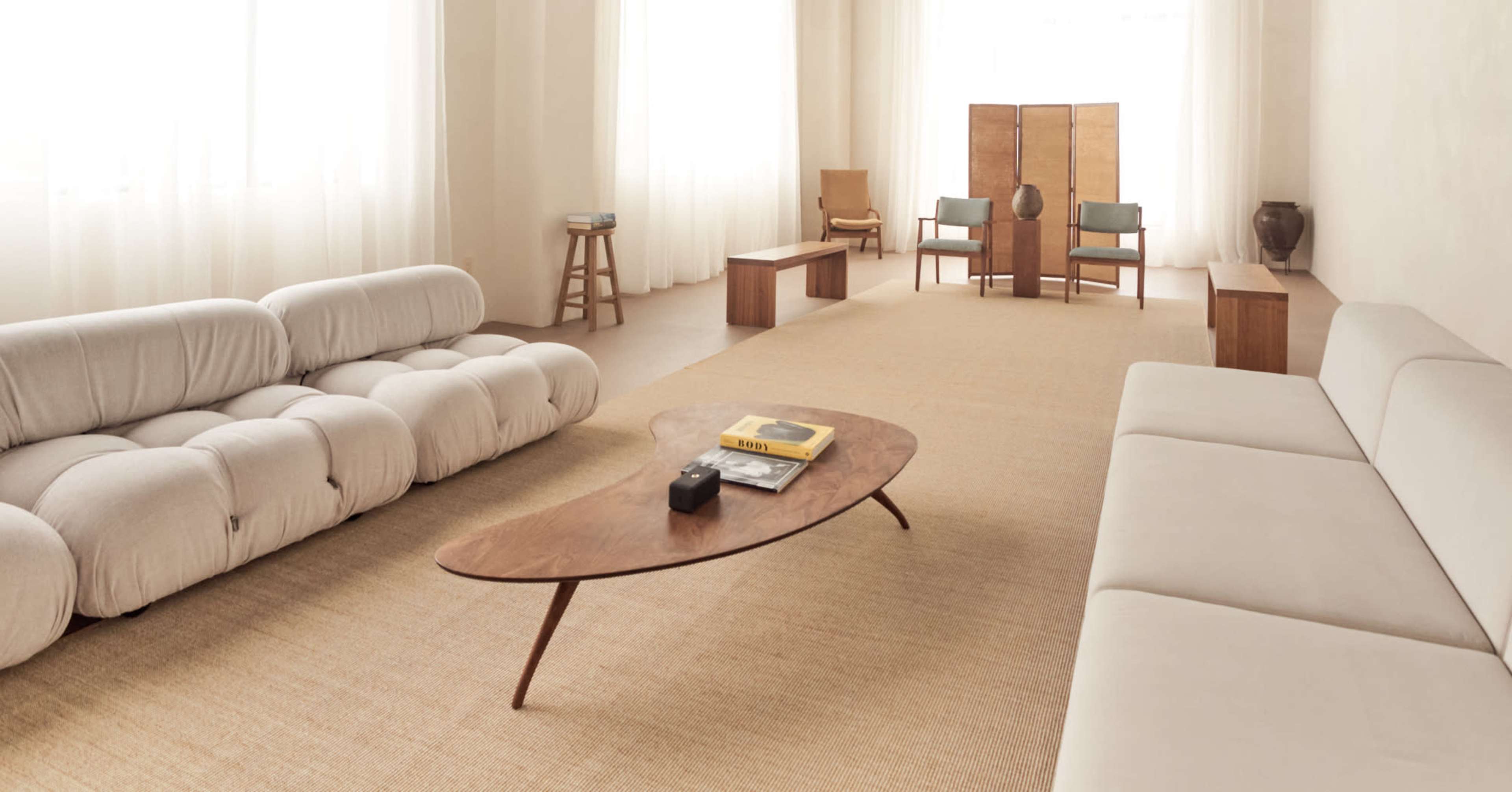 The image depicts a minimalist living room featuring two sofas, a wooden coffee table, and a few chairs arranged around a light-colored area rug.