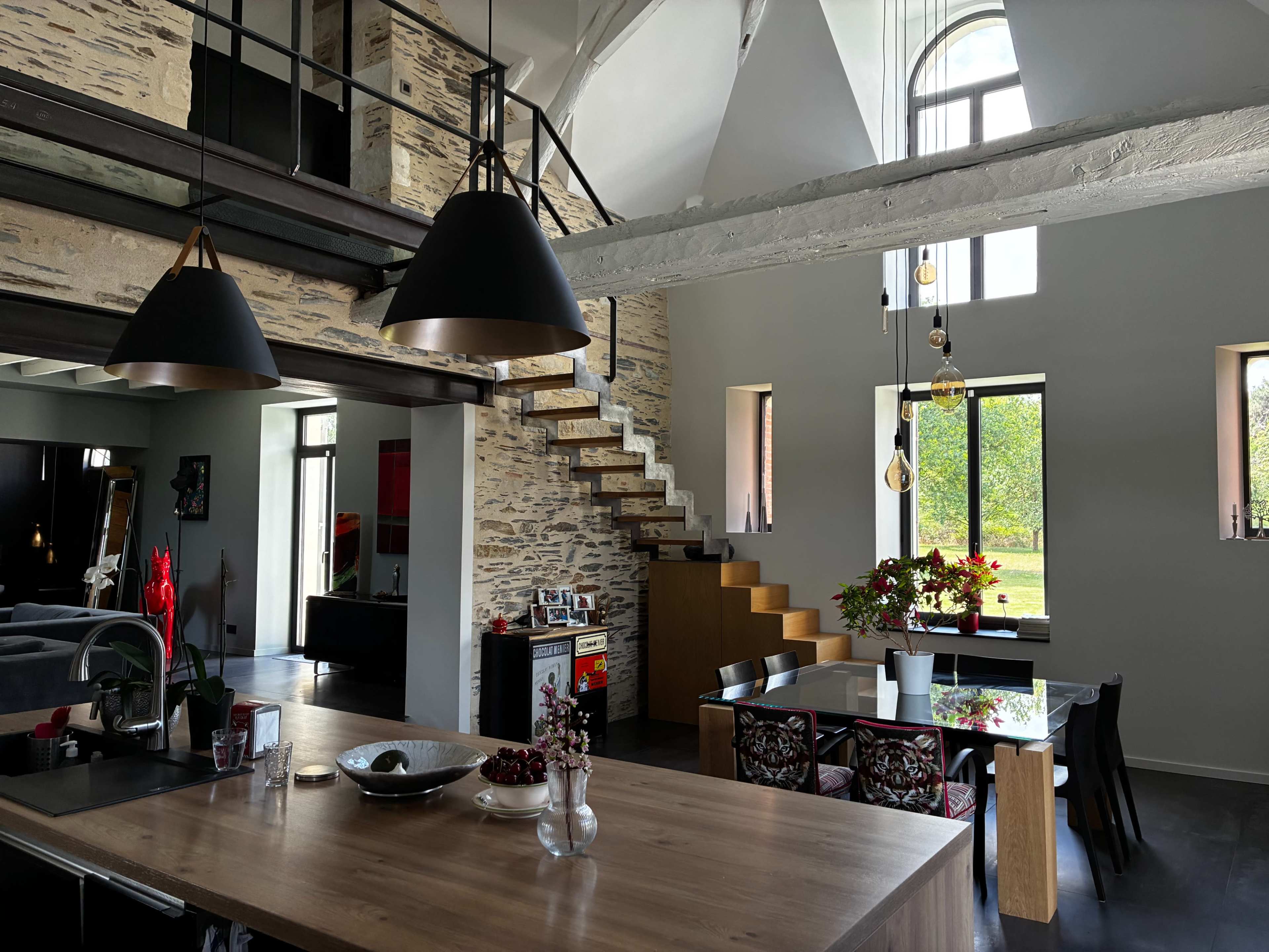 The image shows a modern open-concept living space with a kitchen, dining area, and a staircase leading to a second level, accented by large windows and unique architectural details.