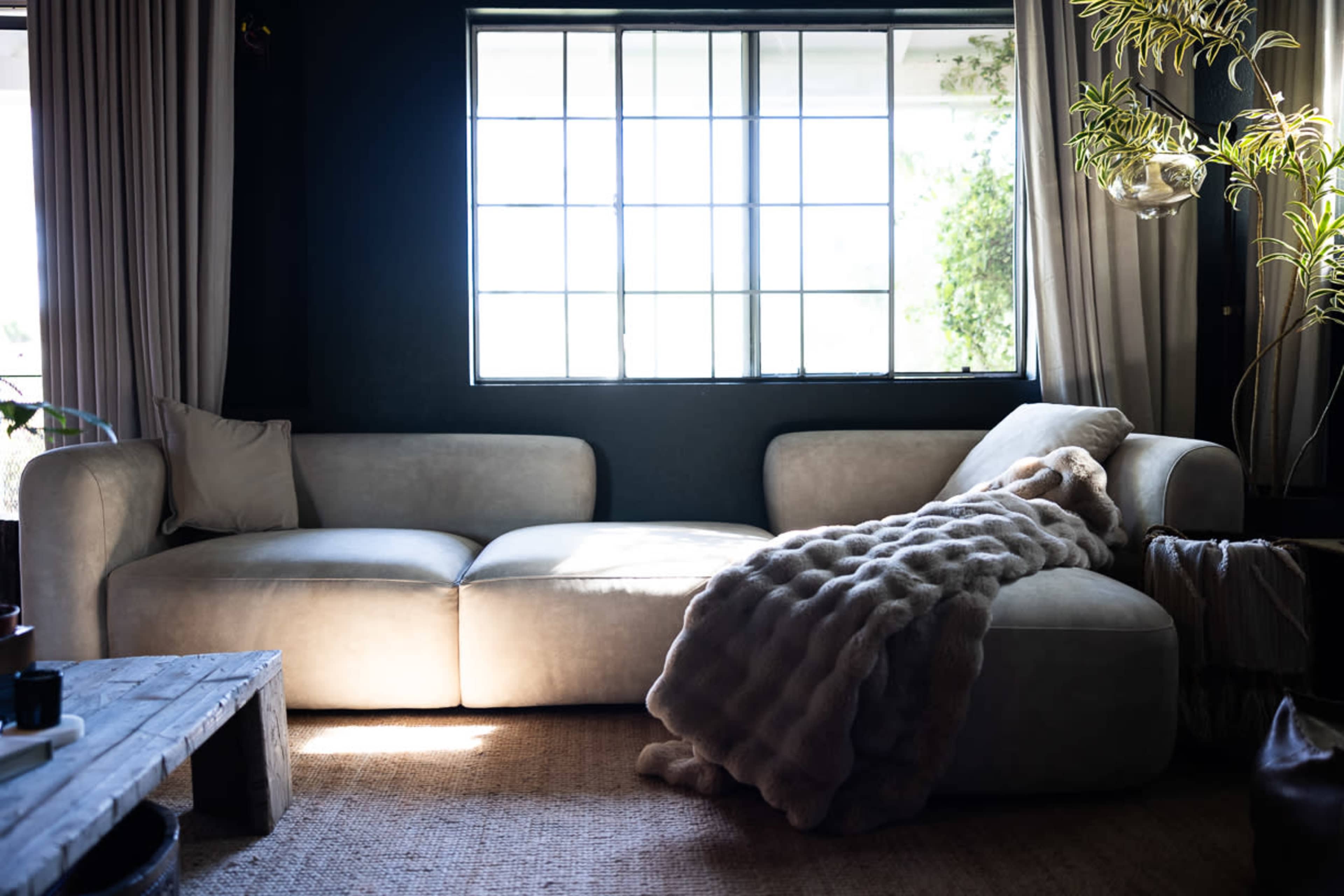 A cozy living room features a light-colored sofa draped with a soft blanket, illuminated by natural light from a large window.