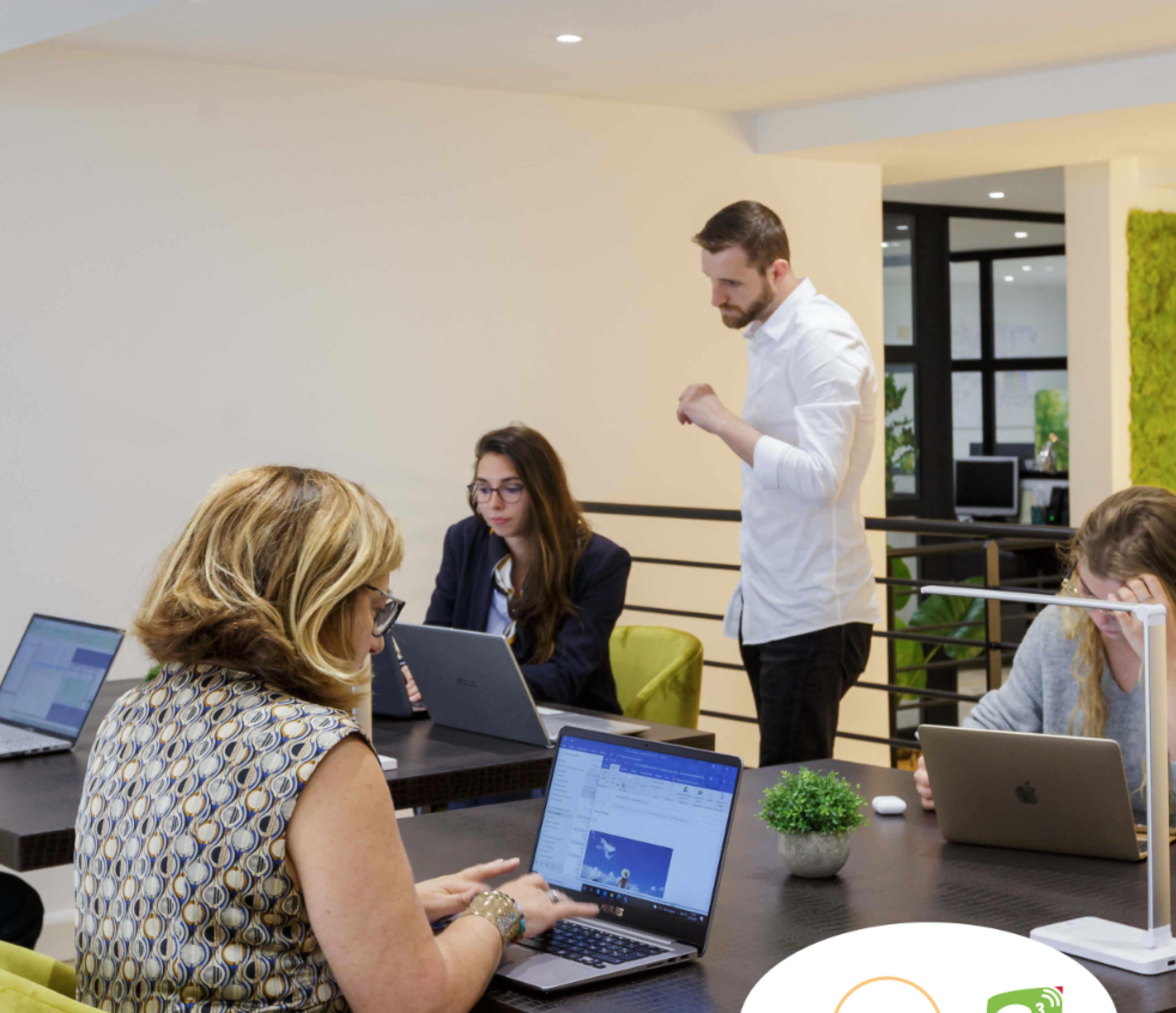 A group of five people is collaborating in a modern office setting, working at laptops while discussing a project.