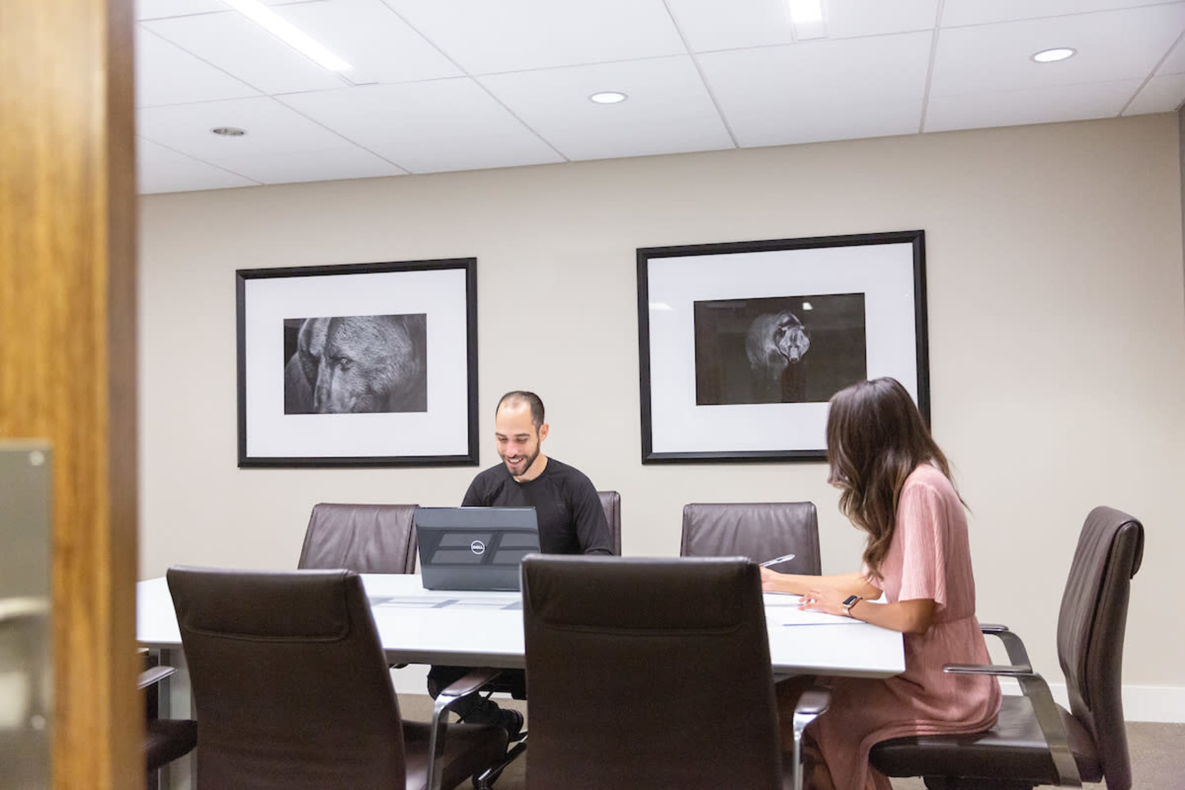 A man sits at a conference table with a laptop, while a woman writes notes next to him in a meeting room featuring two framed black-and-white animal photographs on the walls.