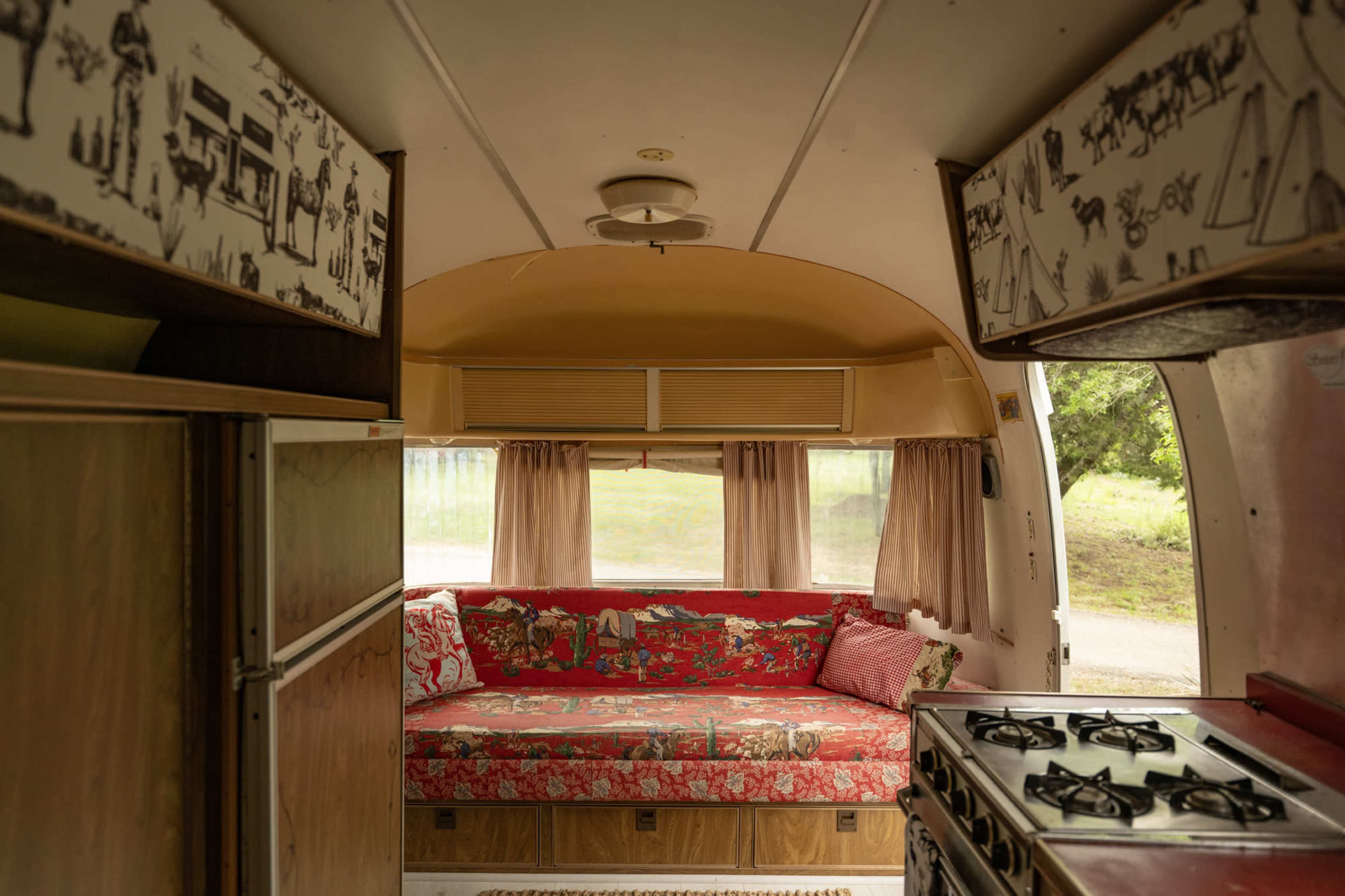 The interior of a vintage camper features a floral sofa, wooden cabinetry, and a kitchen area with a stove.