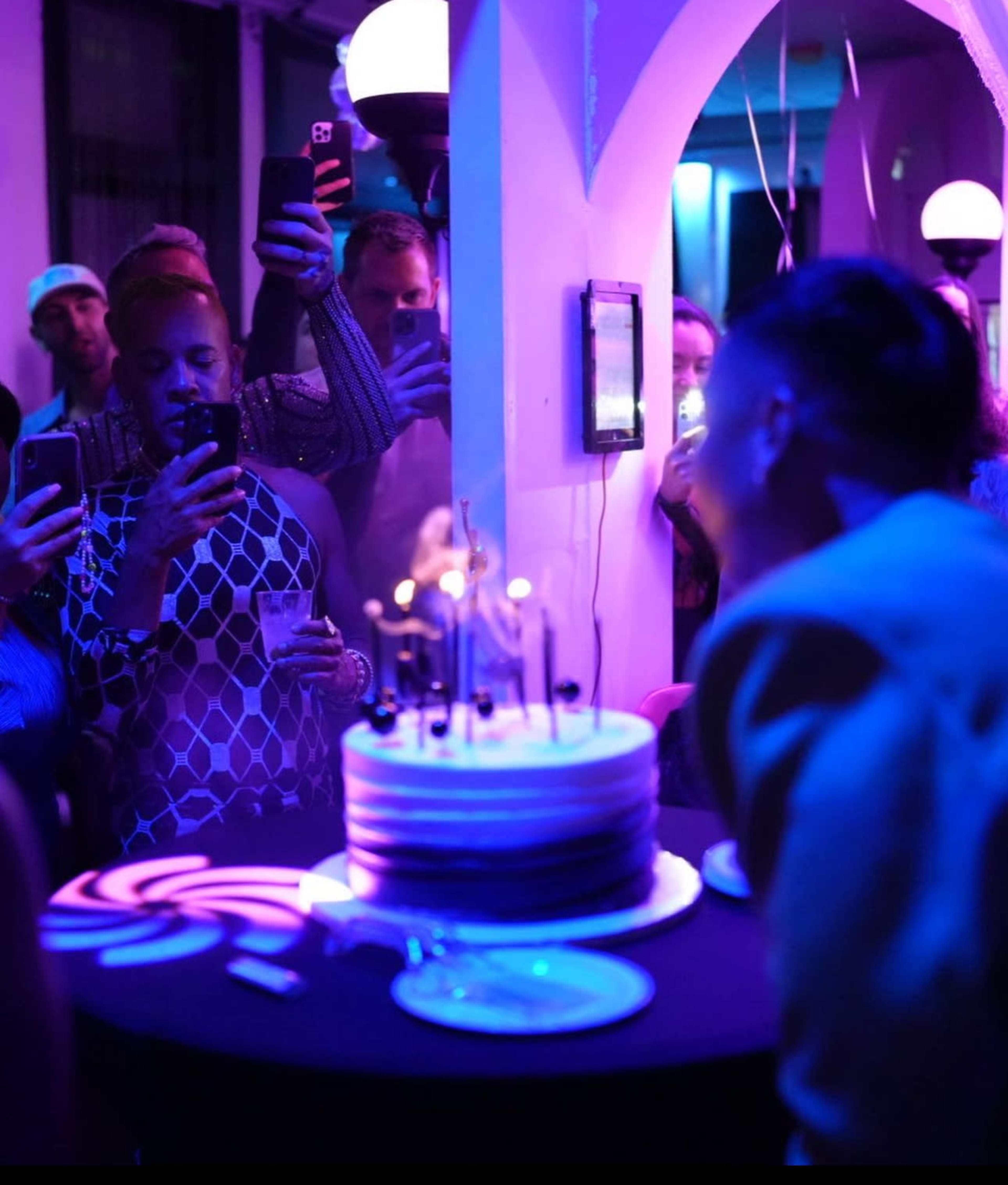 A group of people gathers around a table with a large cake adorned with candles, while several attendees capture the moment on their phones.