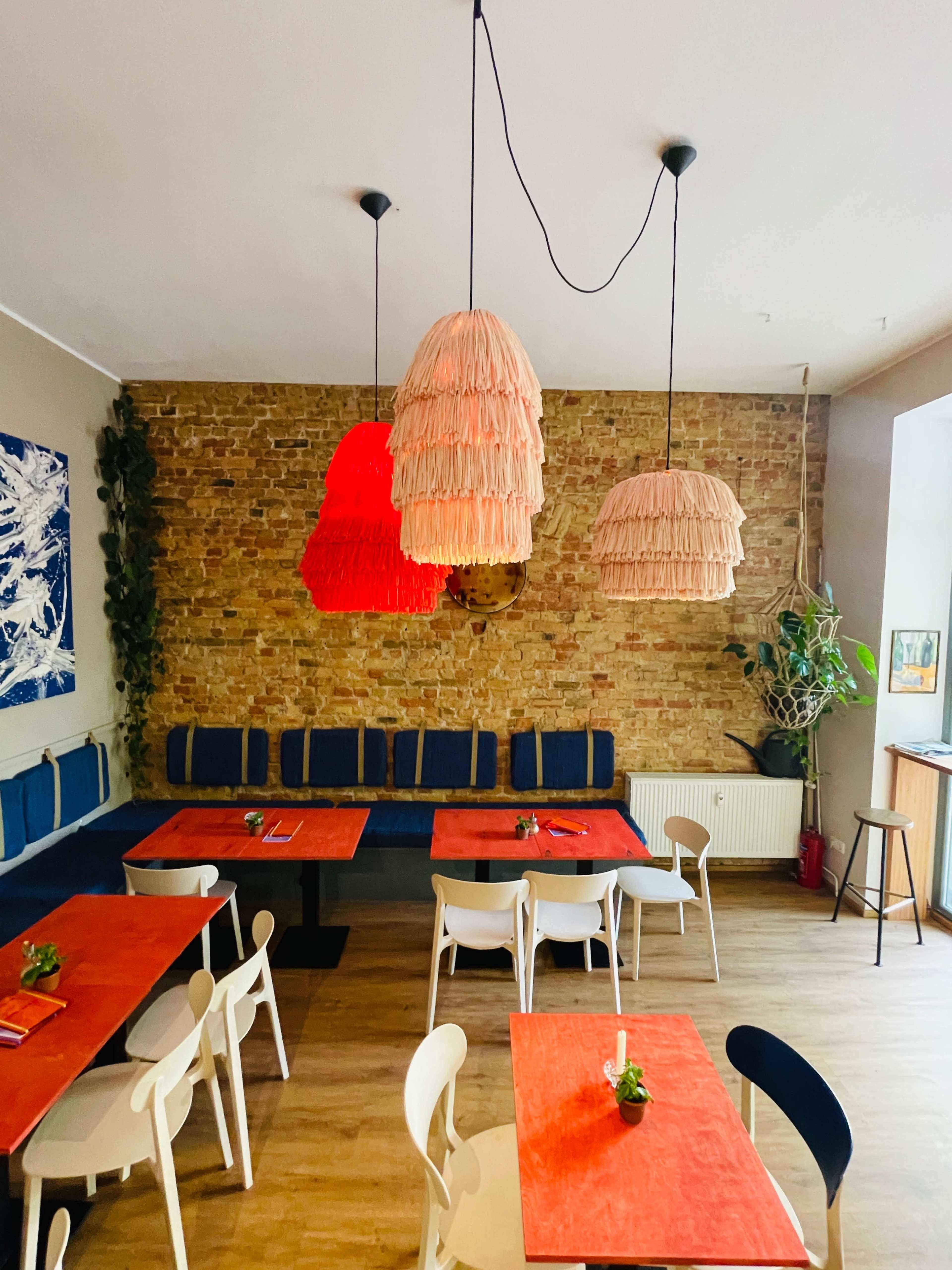 A cozy restaurant features four pendant lamps and red tabletops with white chairs arranged around them, against a brick wall.