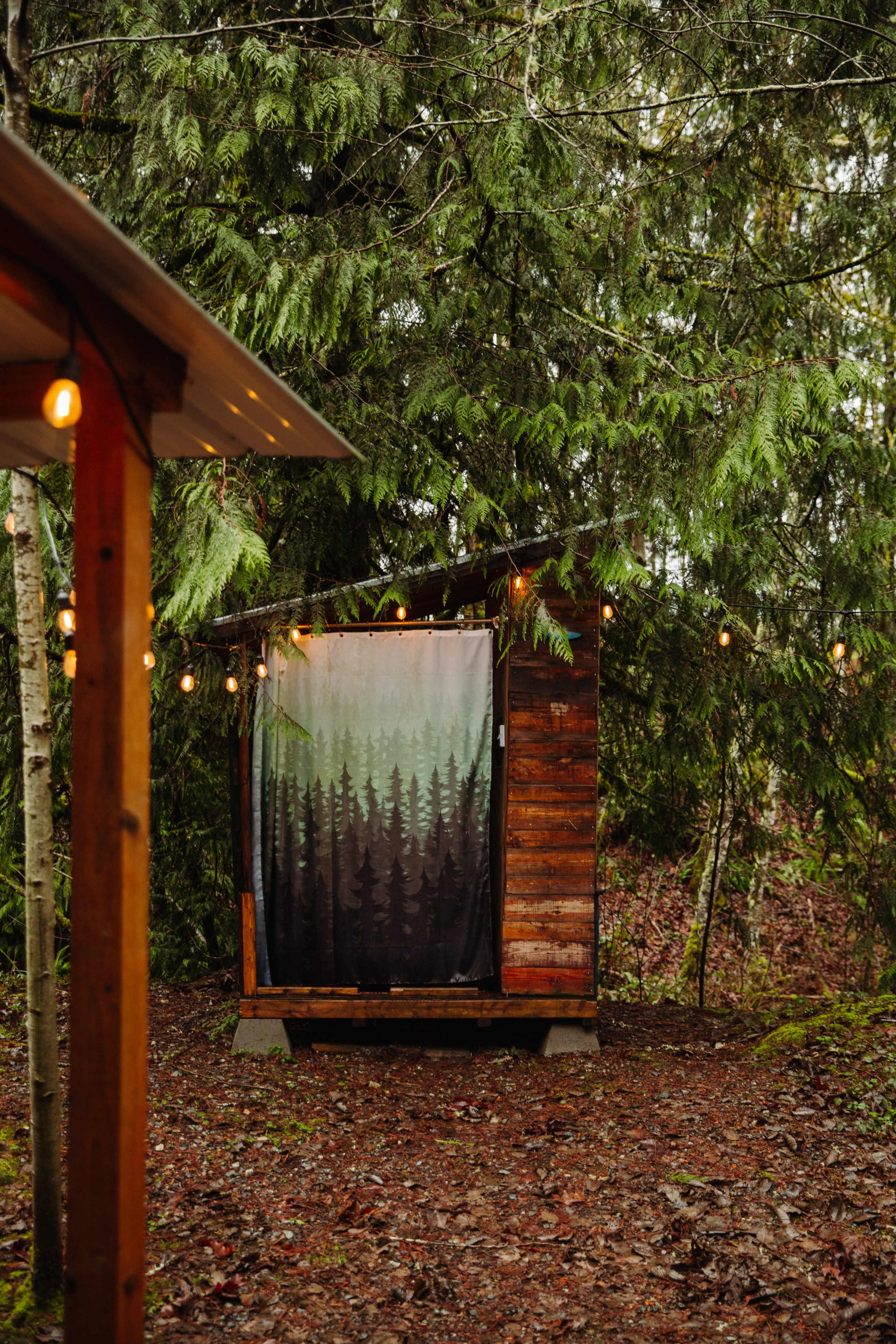 A wooden cabin with a forest-themed curtain is surrounded by tall trees and string lights in a natural setting.