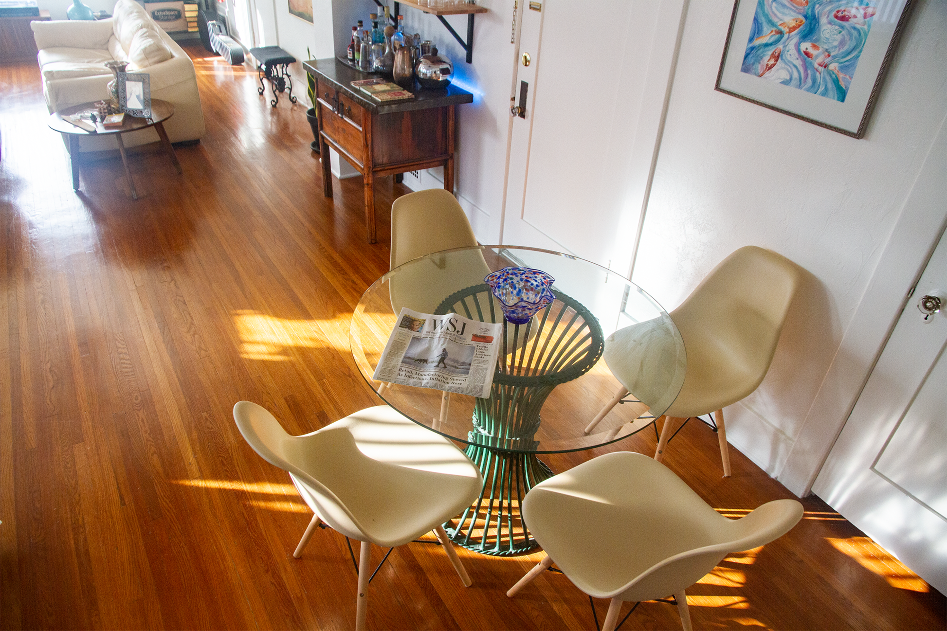A glass dining table surrounded by four white chairs is positioned in a sunlit room with wooden floors and various furnishings in the background.