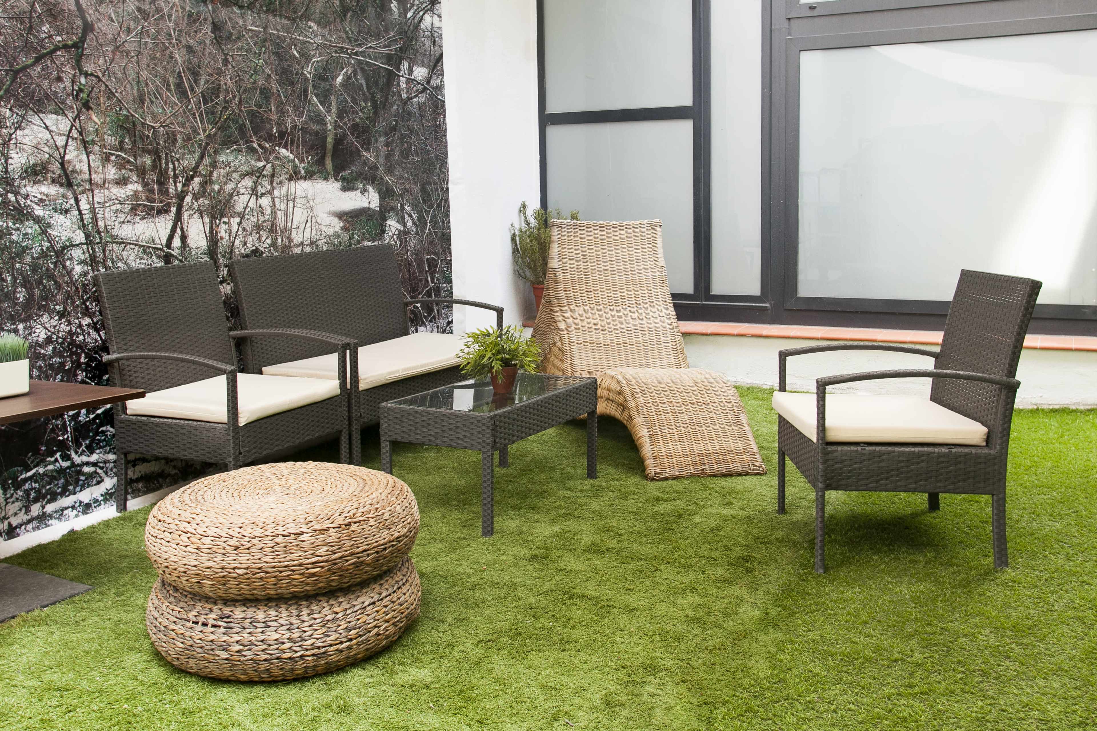 The image shows a patio area furnished with a wicker sofa set, a glass table, a lounge chair, and round woven poufs, all placed on green artificial grass.