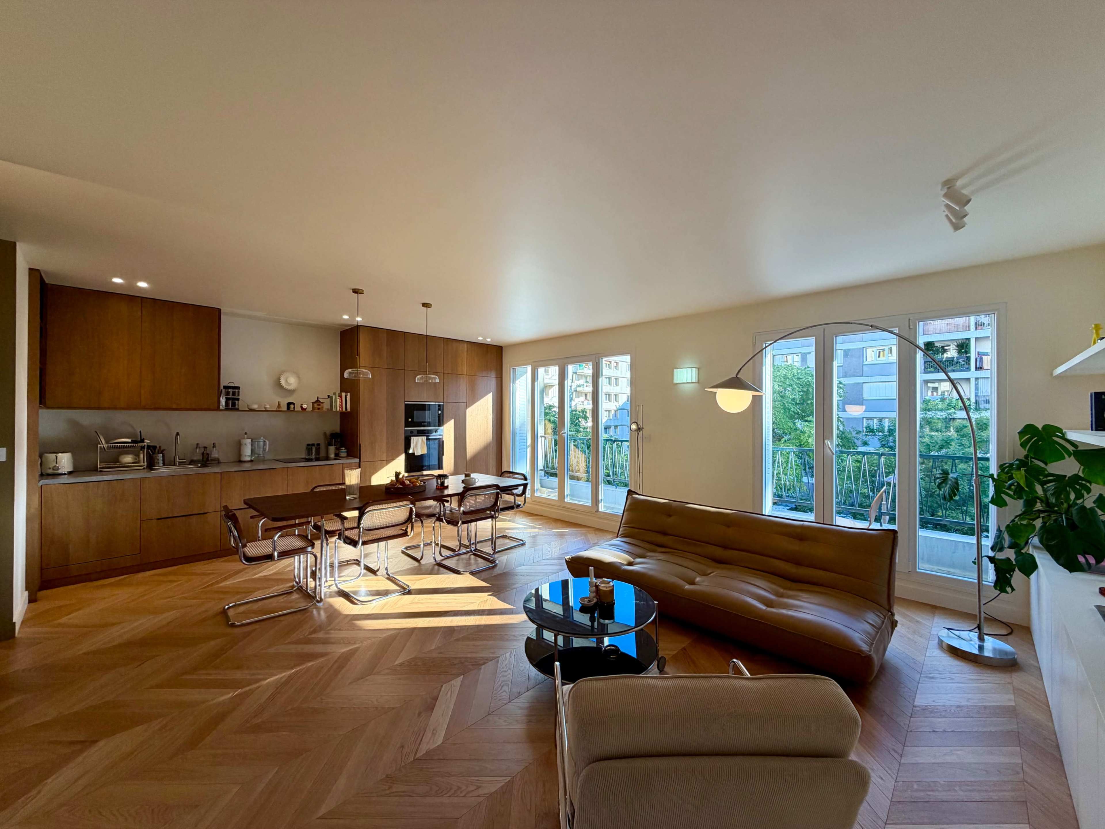 The image shows a modern living space featuring an open layout with a kitchen area, a dining table, a sofa, and large windows that provide natural light and a view of the outdoors.