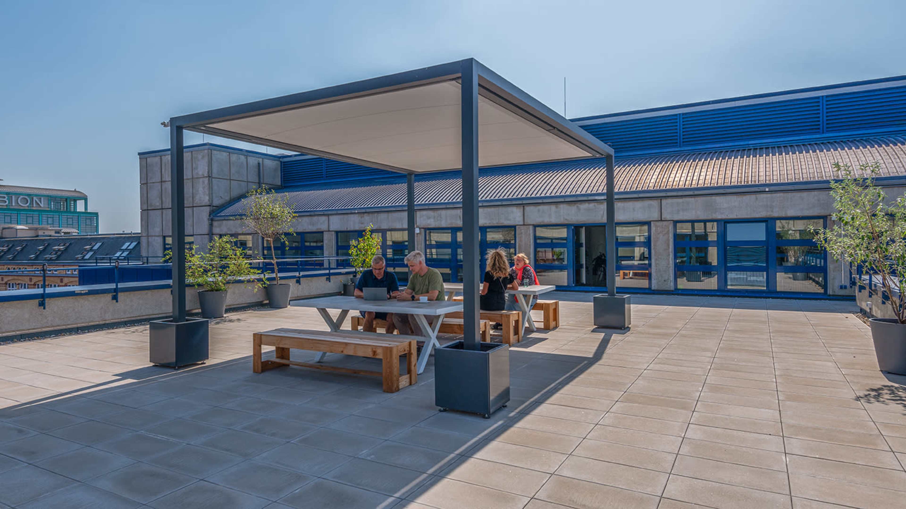 A group of people is sitting at a table under a shaded structure on a rooftop terrace with large windows in the background.