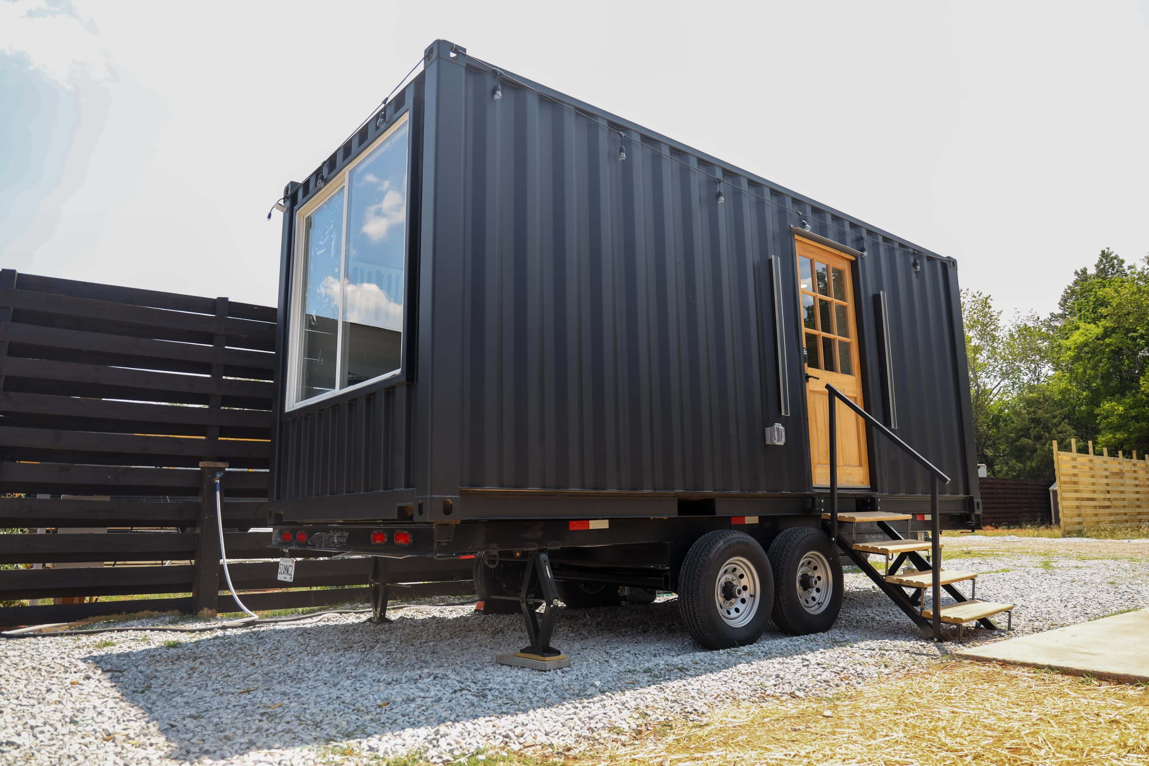 A black shipping container converted into a mobile tiny house is positioned on a gravel surface with a wooden staircase leading to its door.