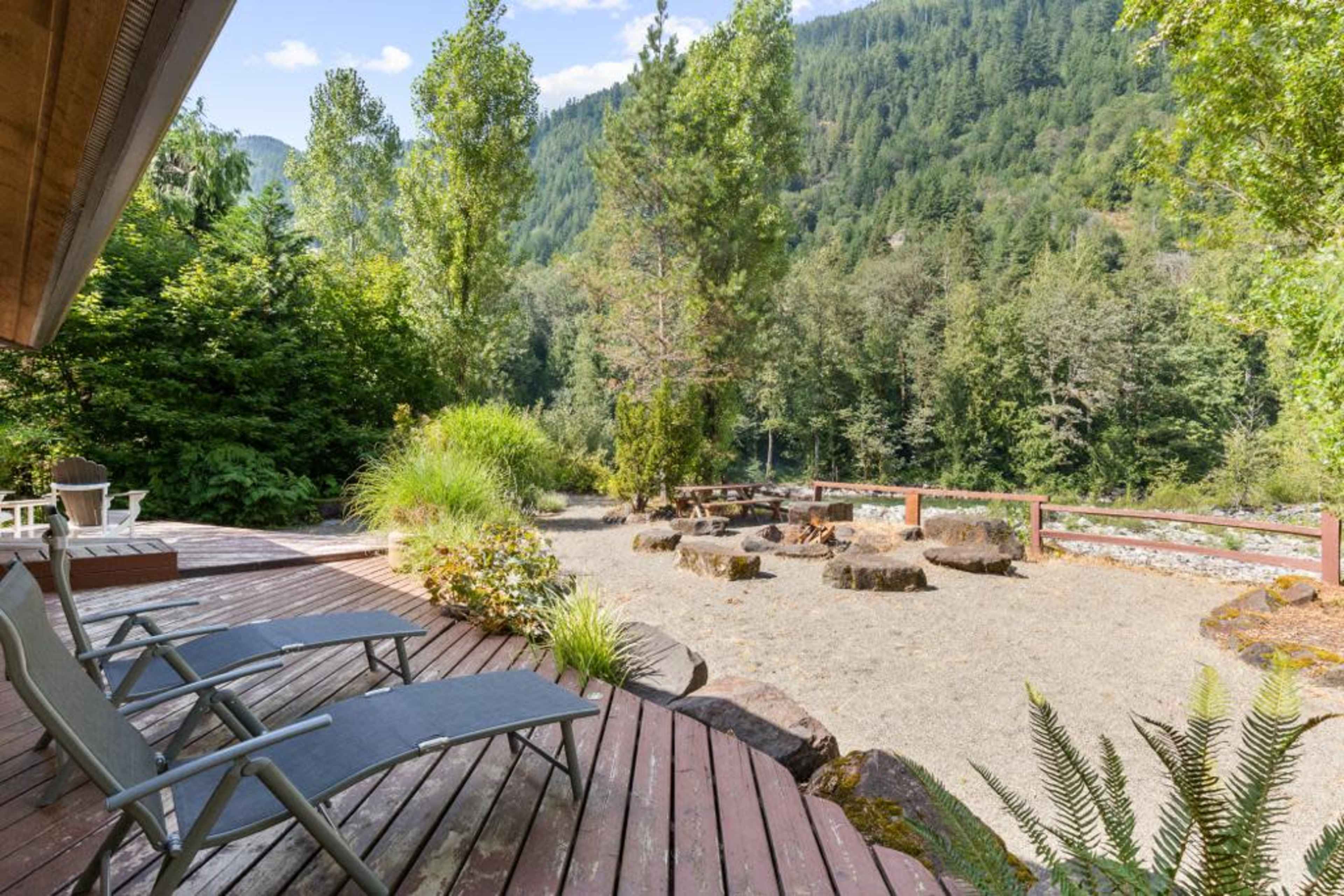 A wooden deck overlooking a landscaped outdoor area with seating logs and a gravel surface surrounded by trees and mountains.
