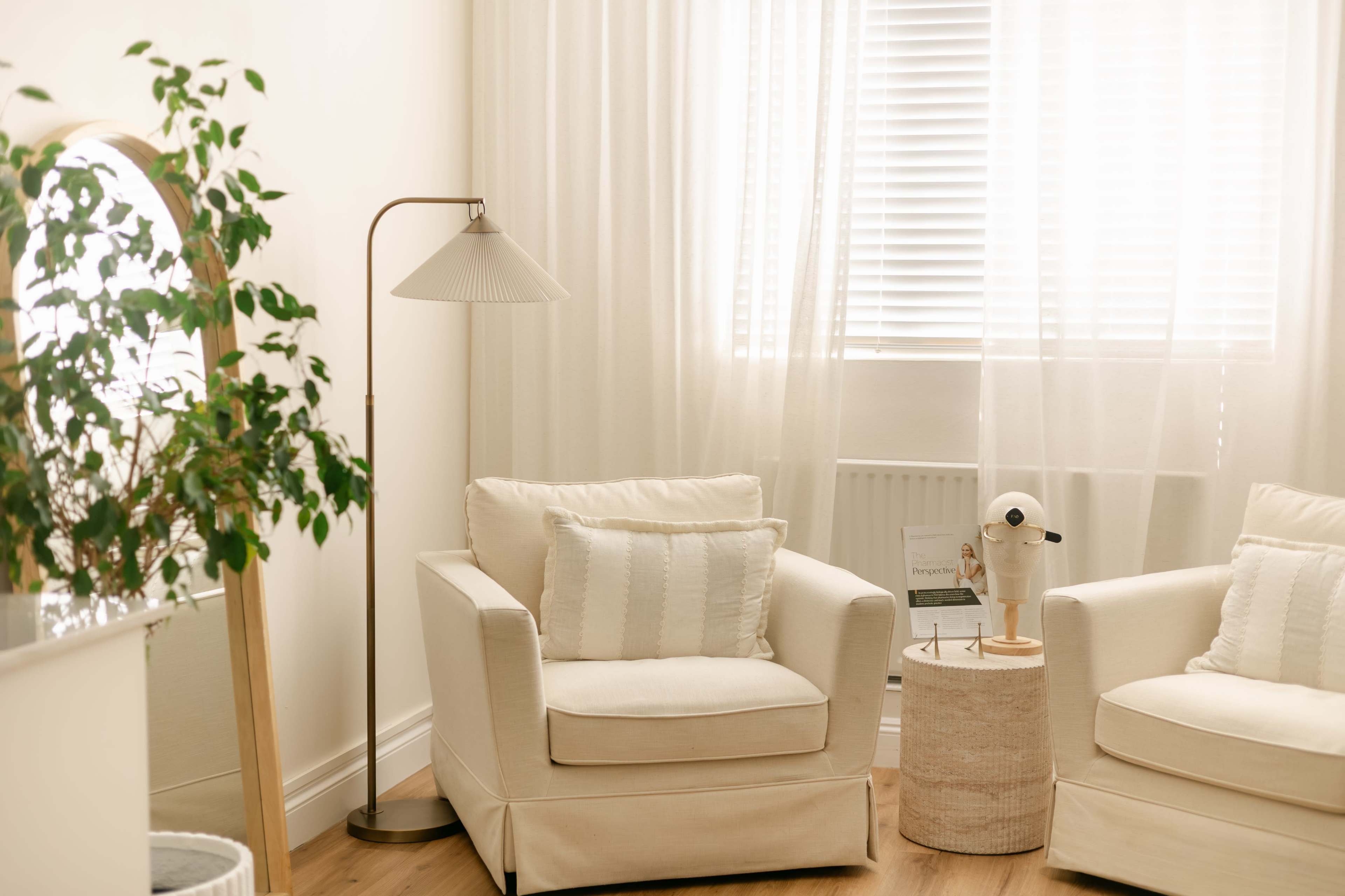 The scene features two white armchairs beside a small round table, with a floor lamp and a potted plant in a bright, softly lit room.