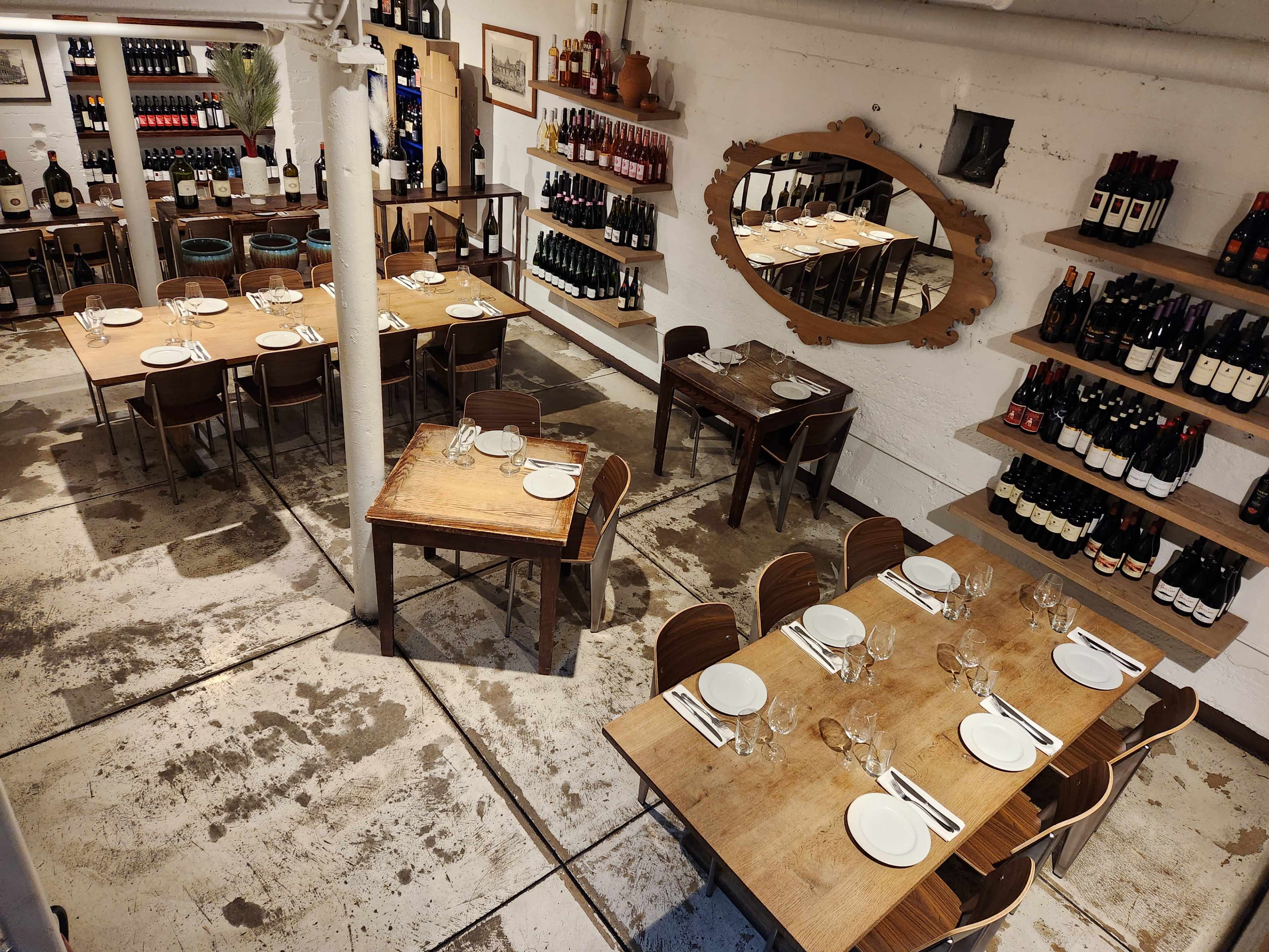 The image shows a restaurant interior with wooden tables arranged in rows, surrounded by shelves filled with wine bottles on the walls.