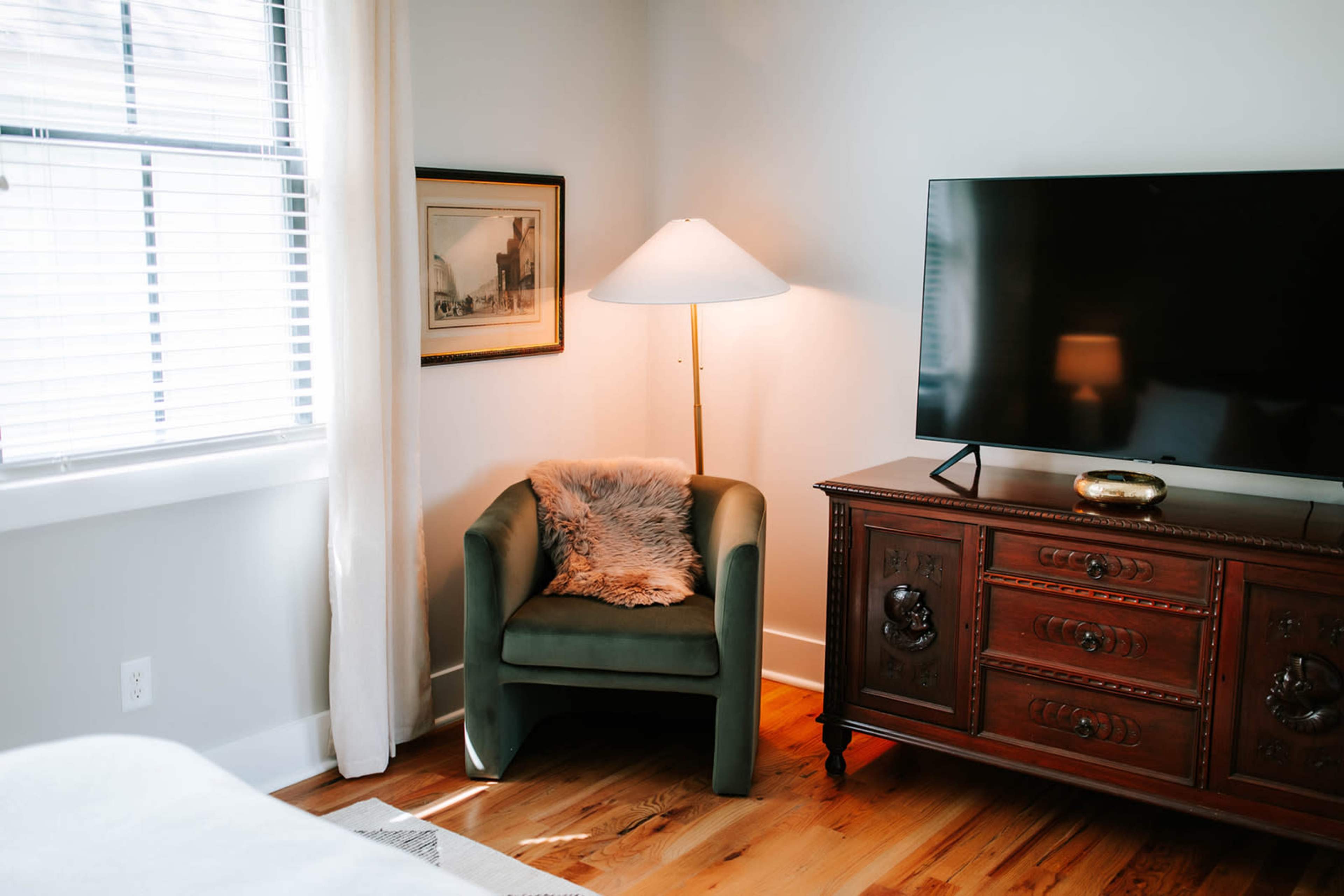 The image shows a cozy room featuring a green armchair with a fur throw, a floor lamp, a wooden dresser with ornate detailing, and a flat-screen TV against a light-colored wall.