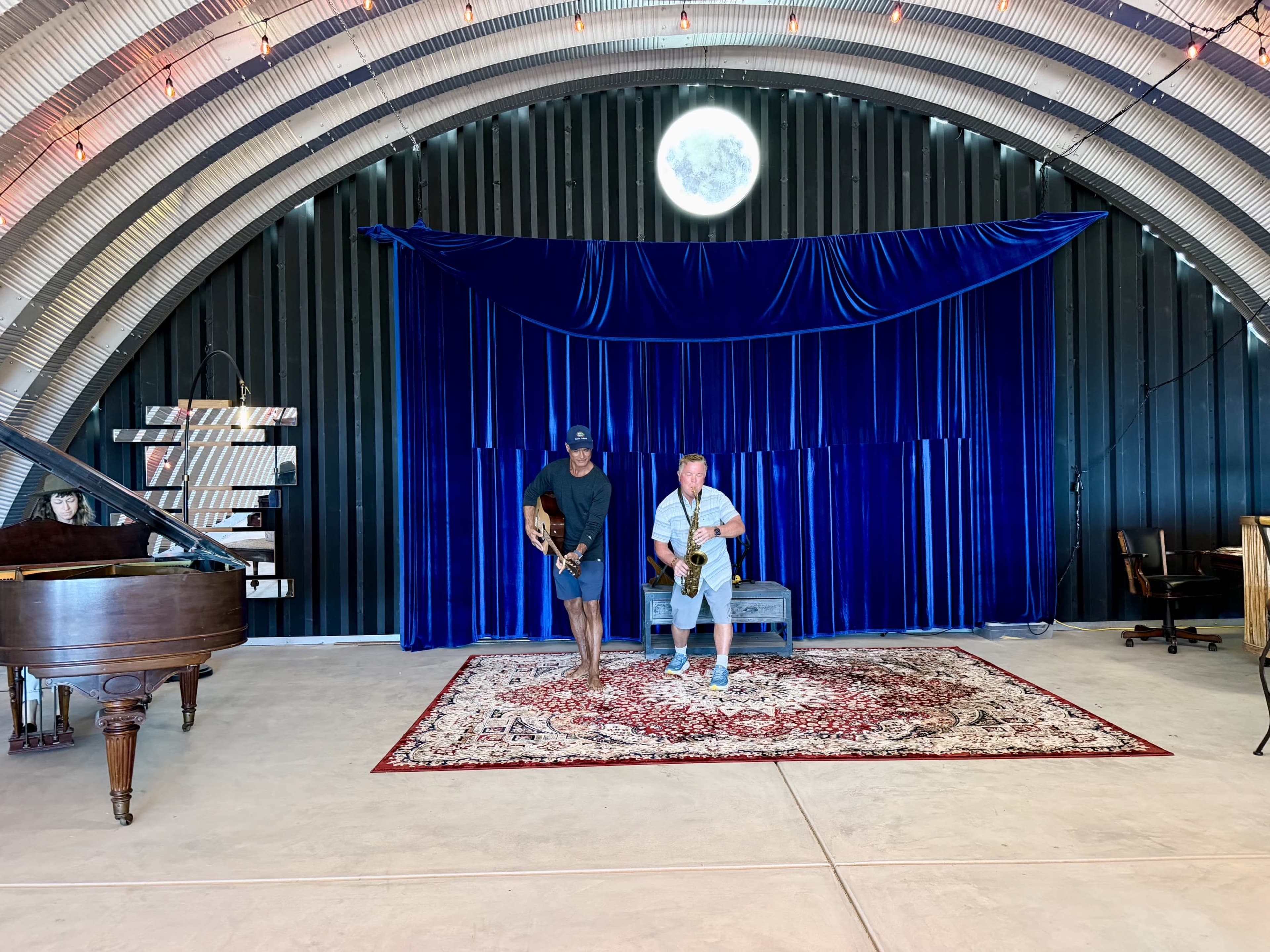Two musicians stand on a patterned rug in front of a blue velvet backdrop inside a venue with a curved metal ceiling and a large circular window.