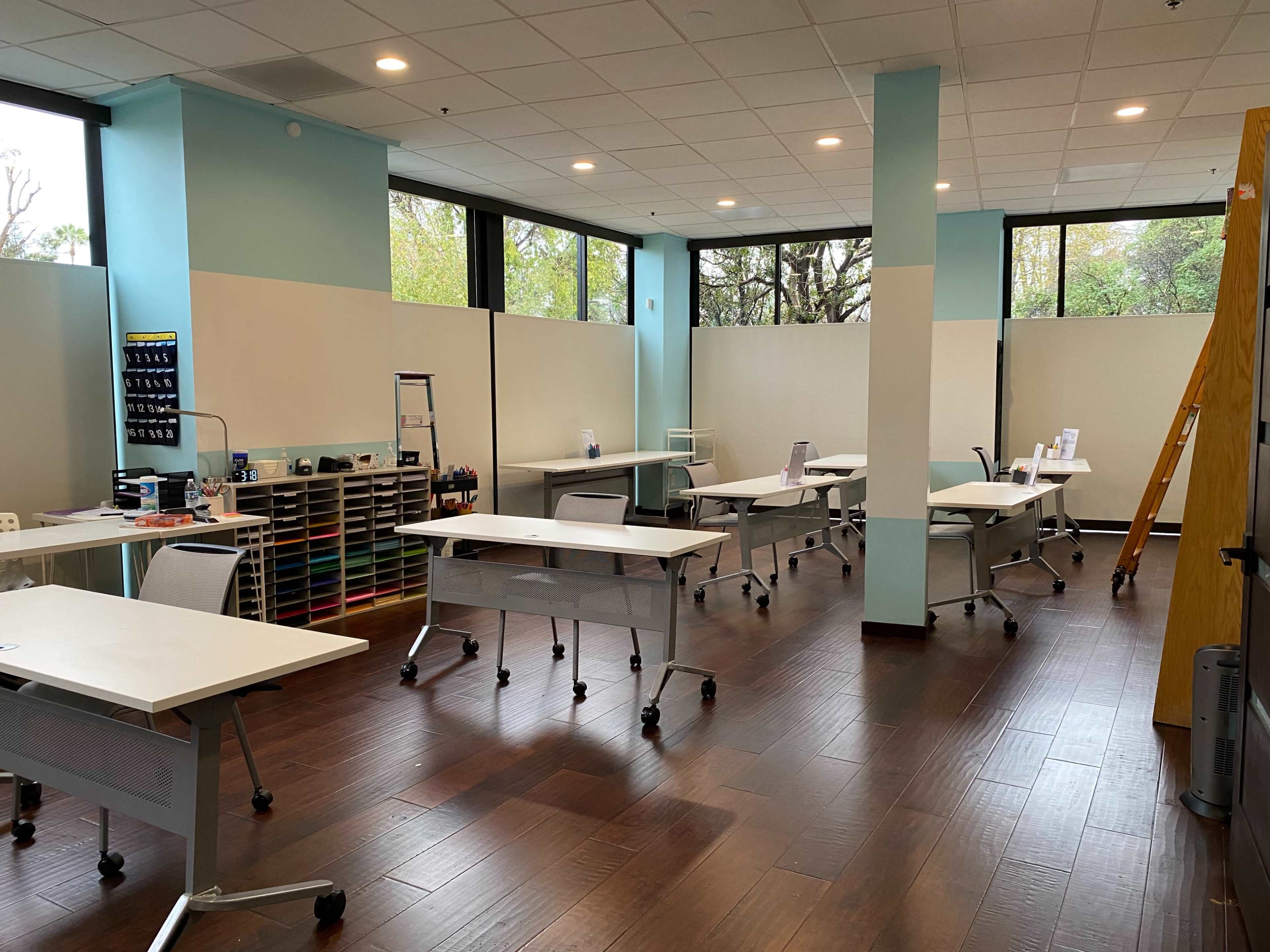 The room features several white desks arranged in a spacious area with large windows and wooden flooring.