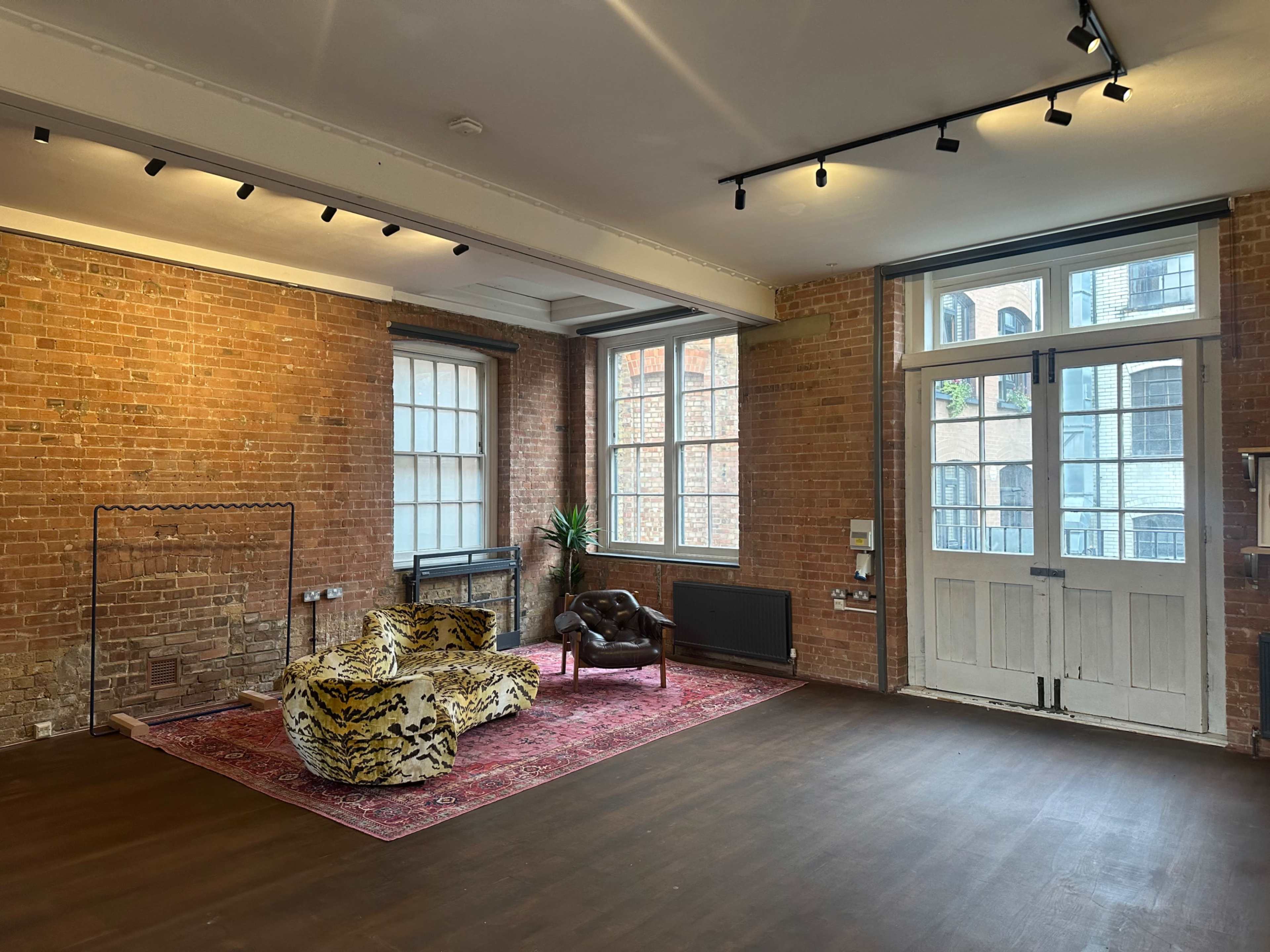 The image shows a spacious room with exposed brick walls, large windows, a patterned sofa, and a vintage chair on a decorative rug.