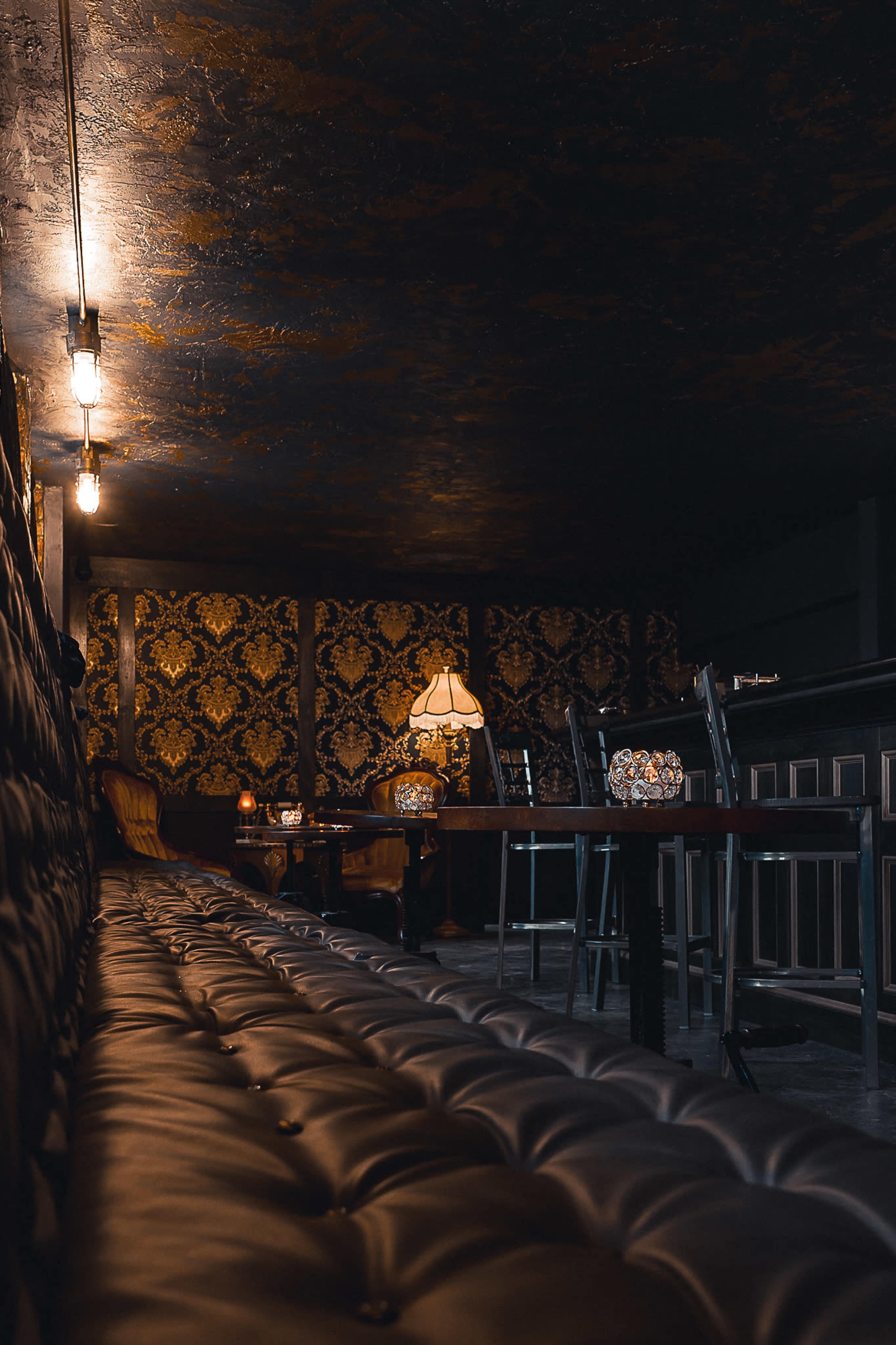 A dimly lit bar with dark, ornate wallpaper, leather seating, and a vintage lamp.