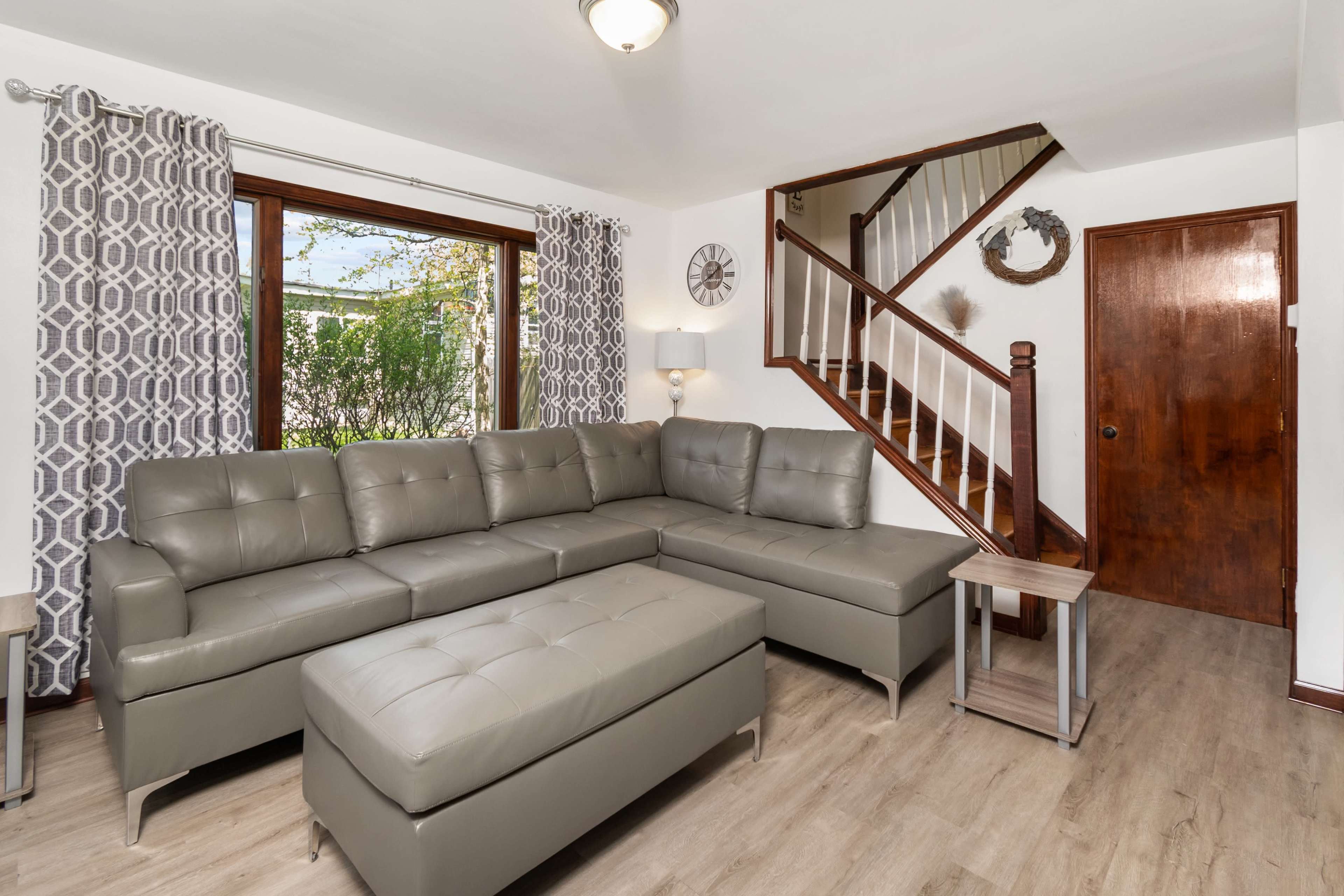 A modern living room features a gray sectional sofa, a matching ottoman, a wooden side table, and a staircase in the corner.