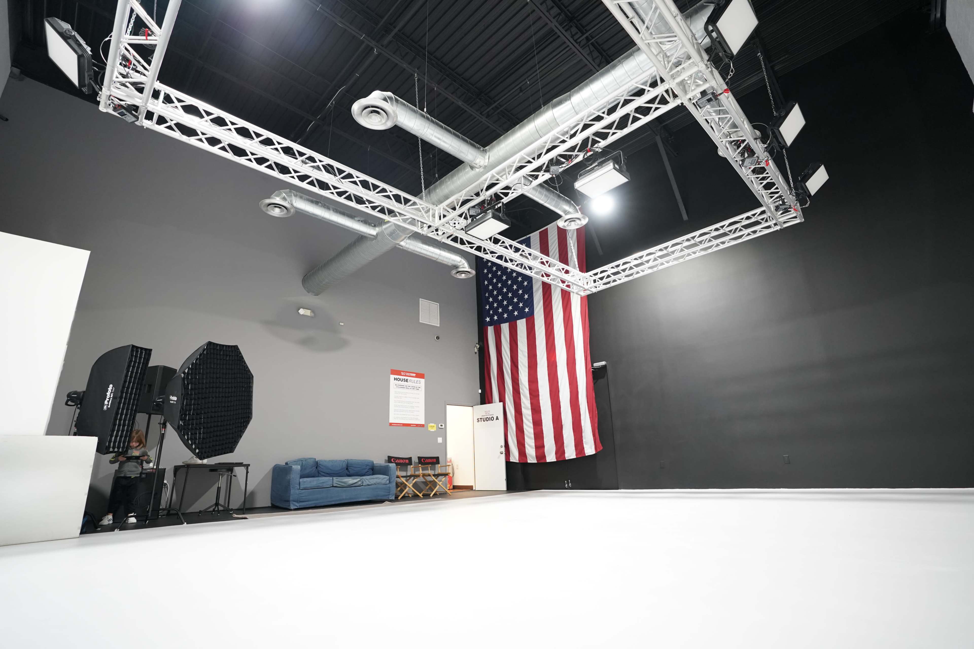 The image shows a photography studio with a white floor, a large American flag hanging on the wall, and various lighting equipment overhead.