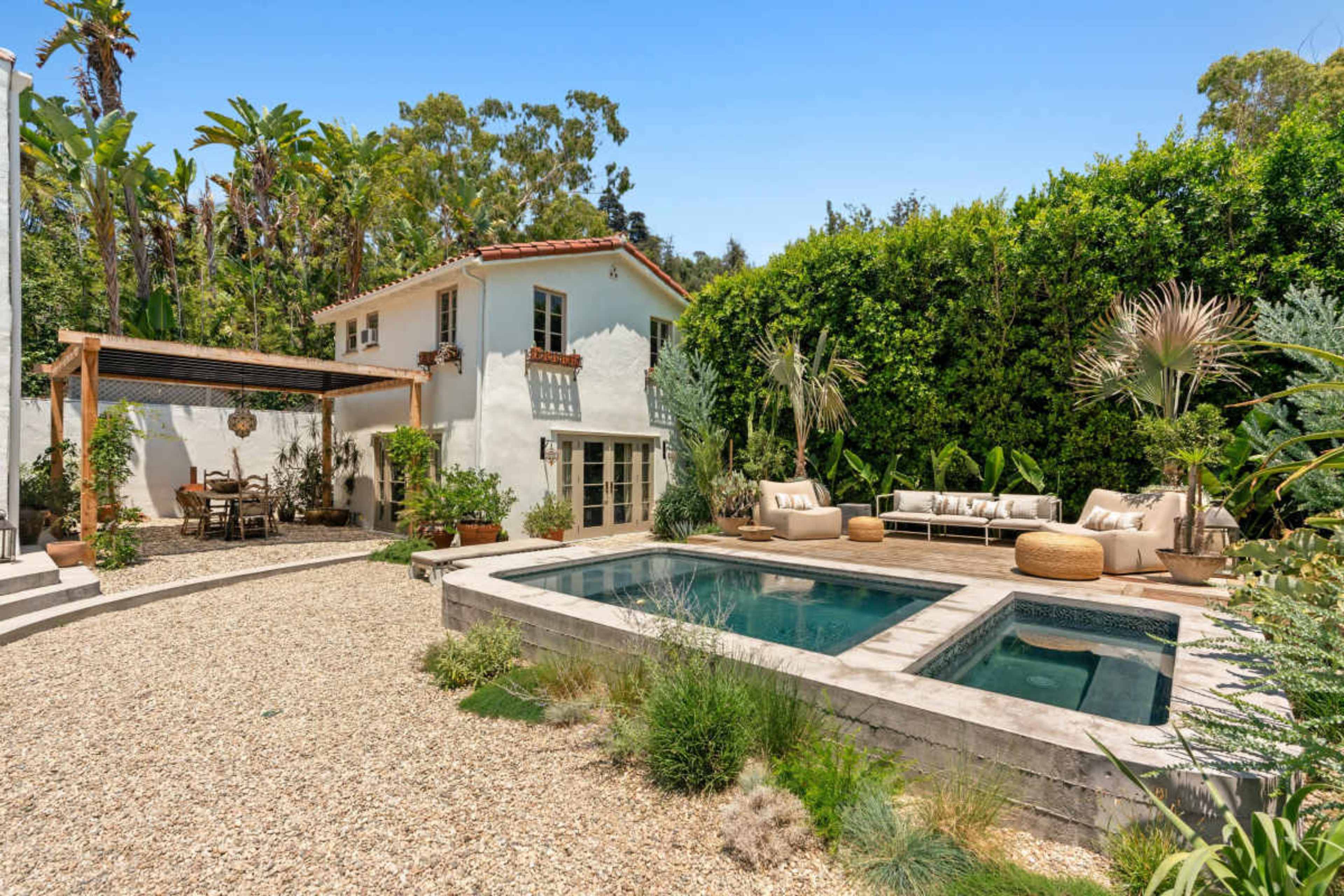 The image shows a landscaped outdoor space featuring a swimming pool, a patio area with seating, and a two-story white house surrounded by greenery.