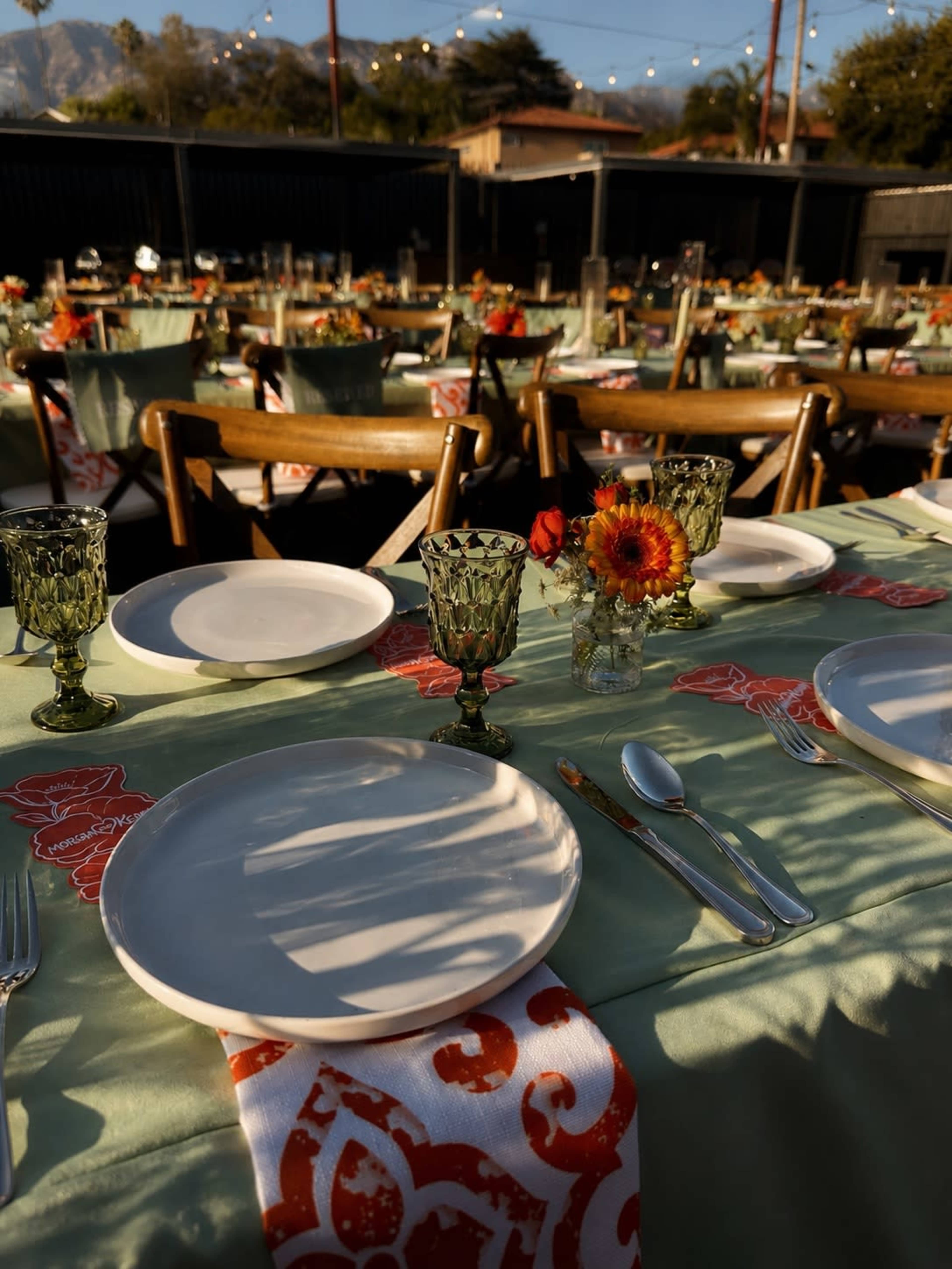 The image shows a beautifully set outdoor dining area with tables adorned with green tablecloths, decorative napkins, and floral centerpieces.