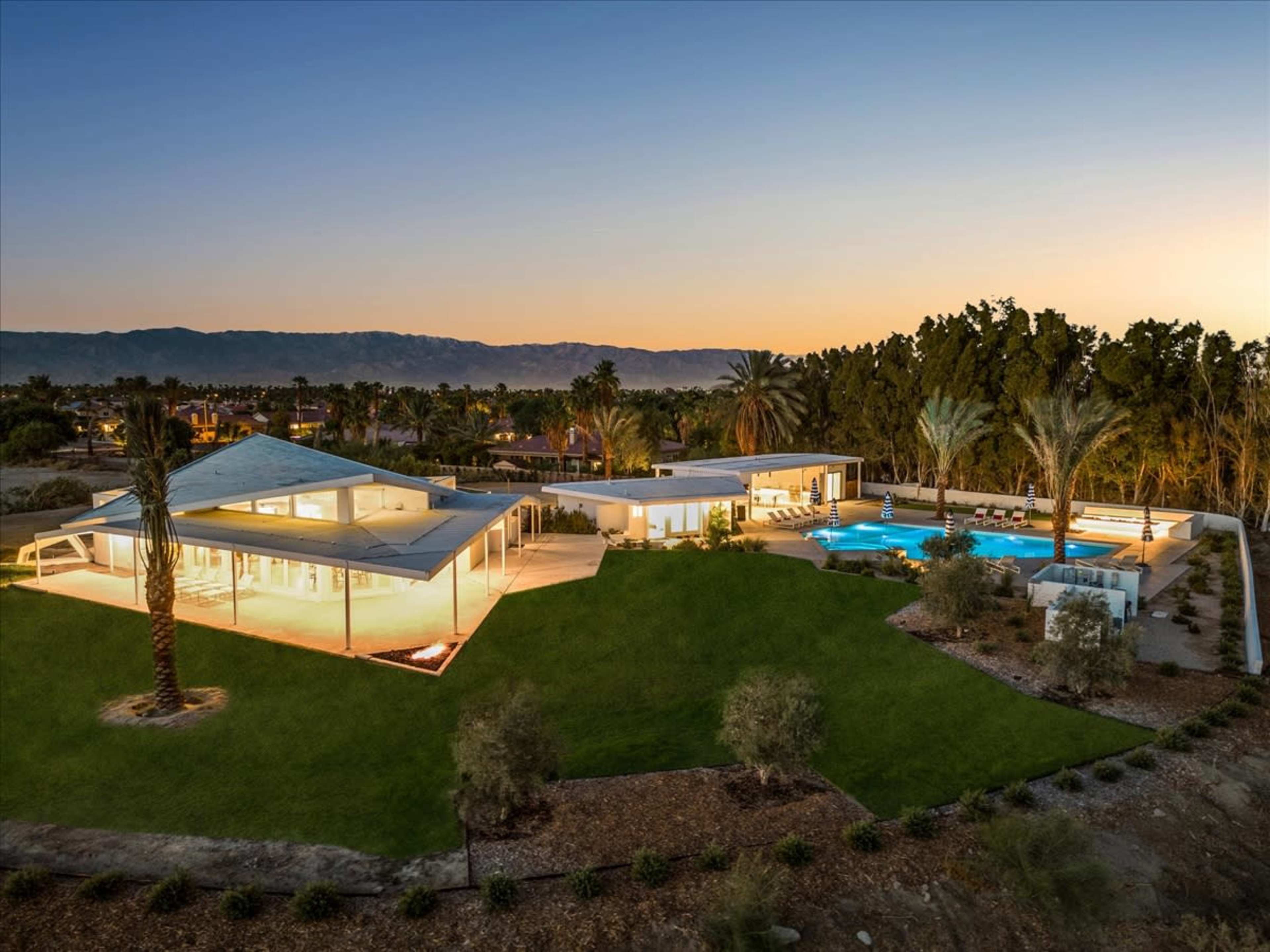 A modern single-story house with lighted patios and a pool, surrounded by landscaped greenery and mountains in the background at sunset.