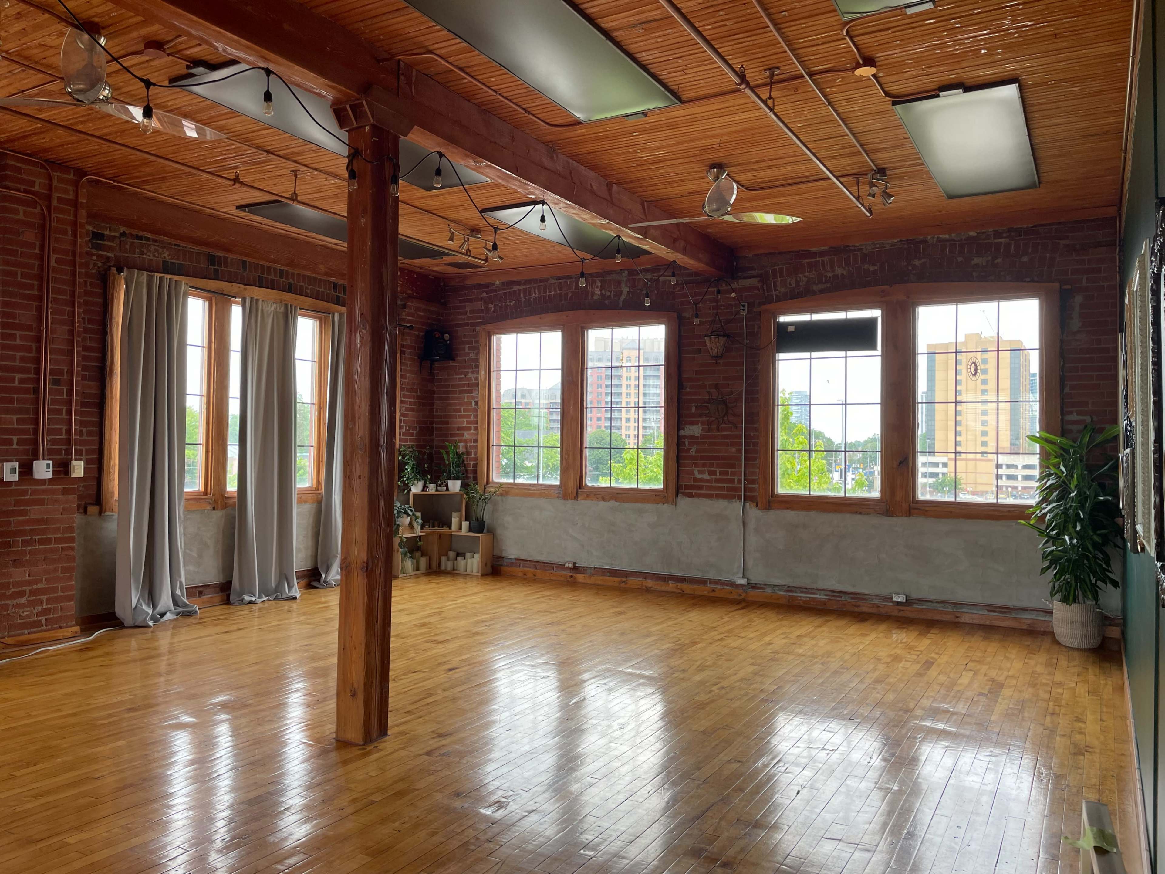 The image shows a spacious room with wooden floors, exposed brick walls, large windows, and natural light coming in.