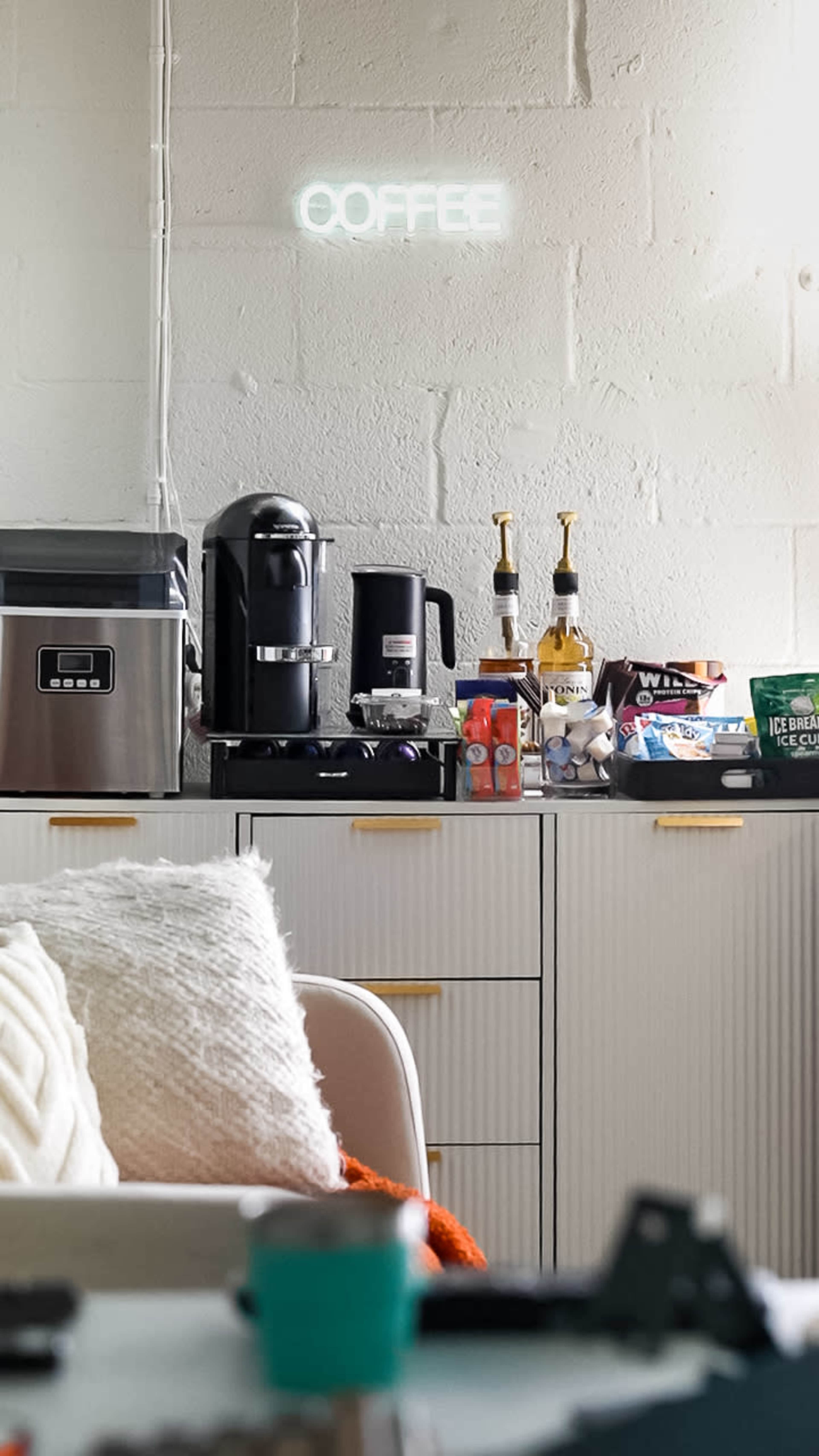 The image shows a kitchen area with a coffee maker, syrups, and various snacks on a countertop beneath a glowing "COFFEE" sign.