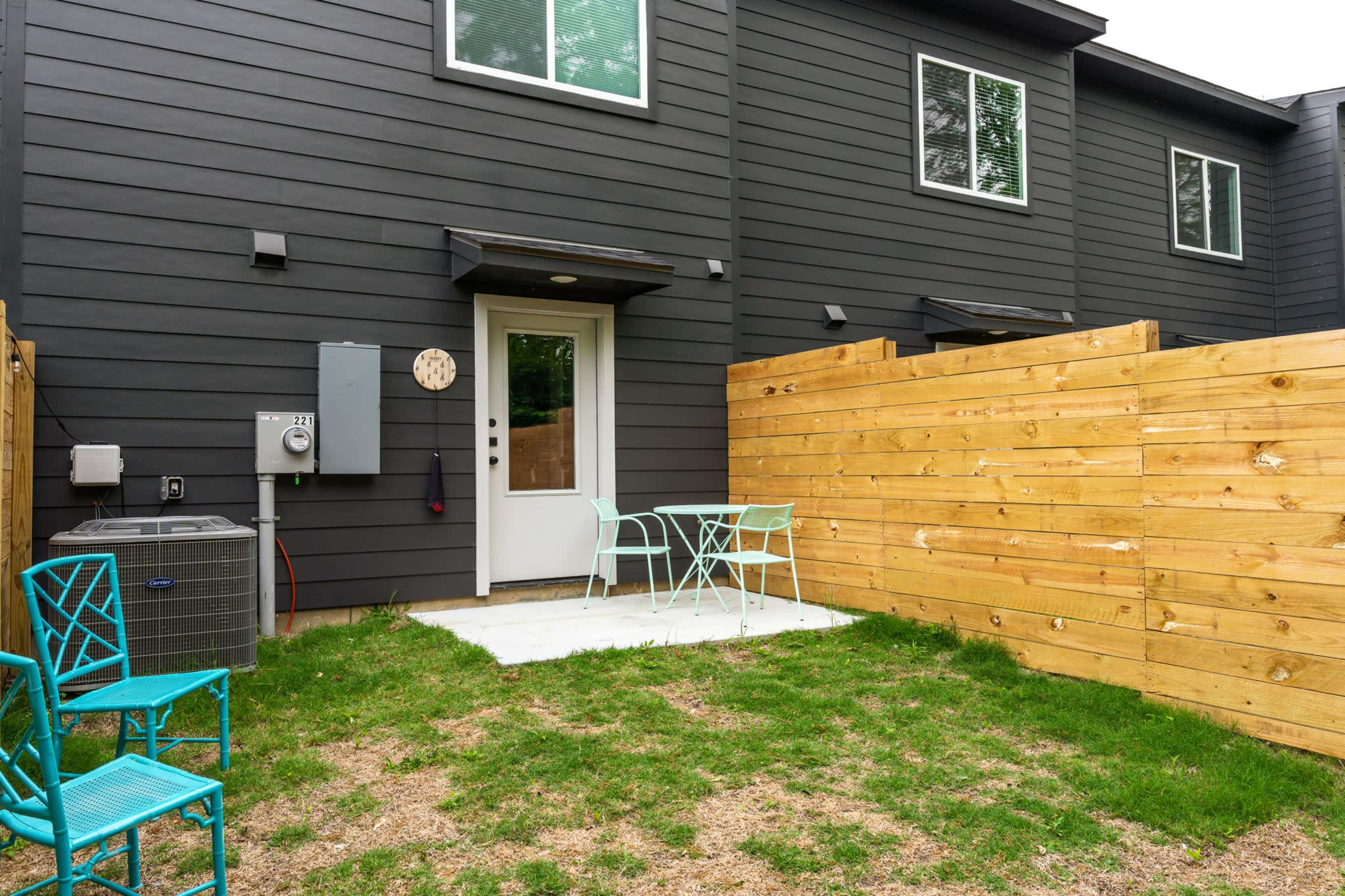 A small outdoor patio area with a light-colored table and two blue chairs surrounded by a wooden fence and grass.