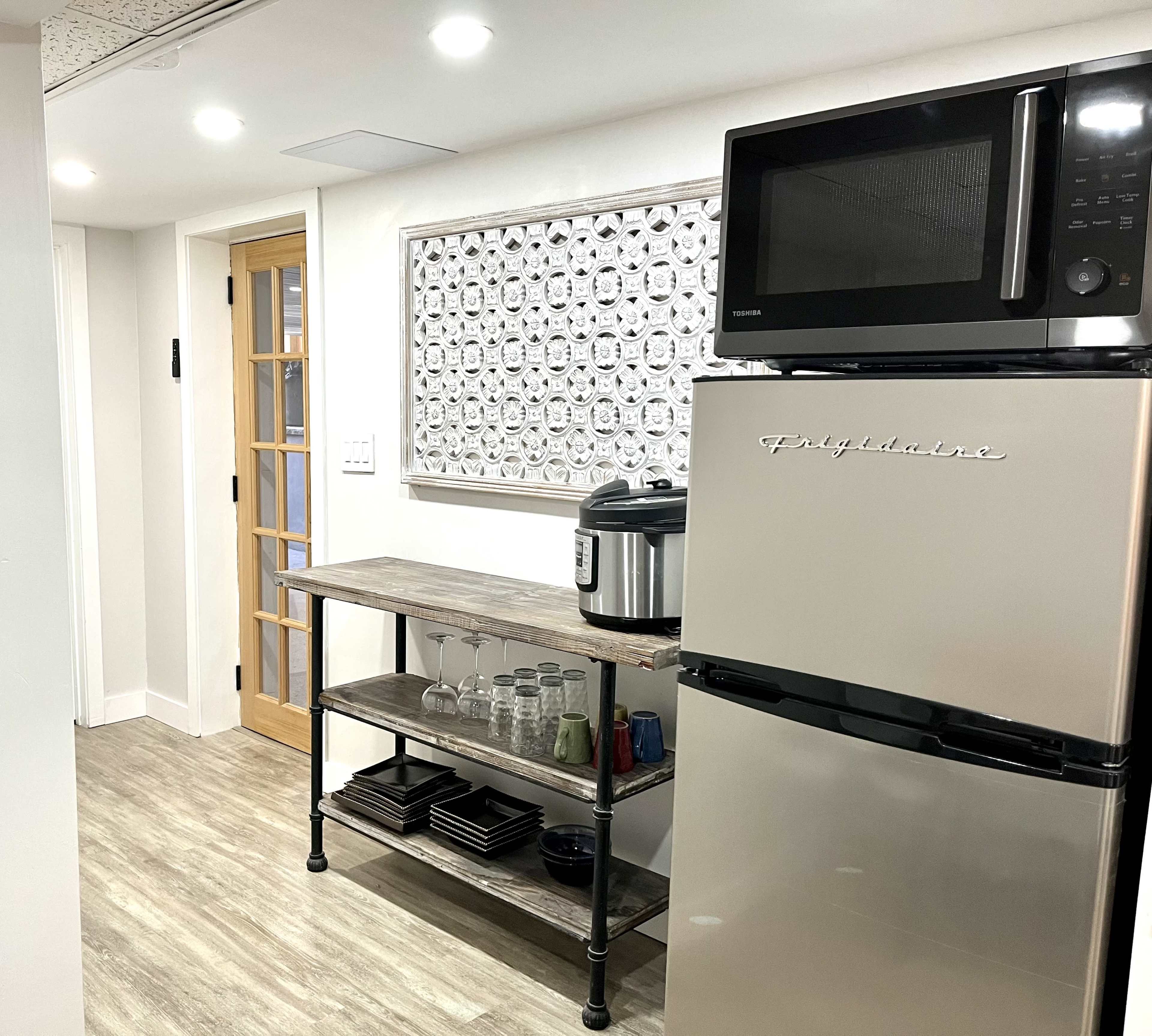 A modern kitchen with a stainless steel refrigerator, a microwave above it, a wooden shelf holding glassware and dishes, and a doorway leading to another room.