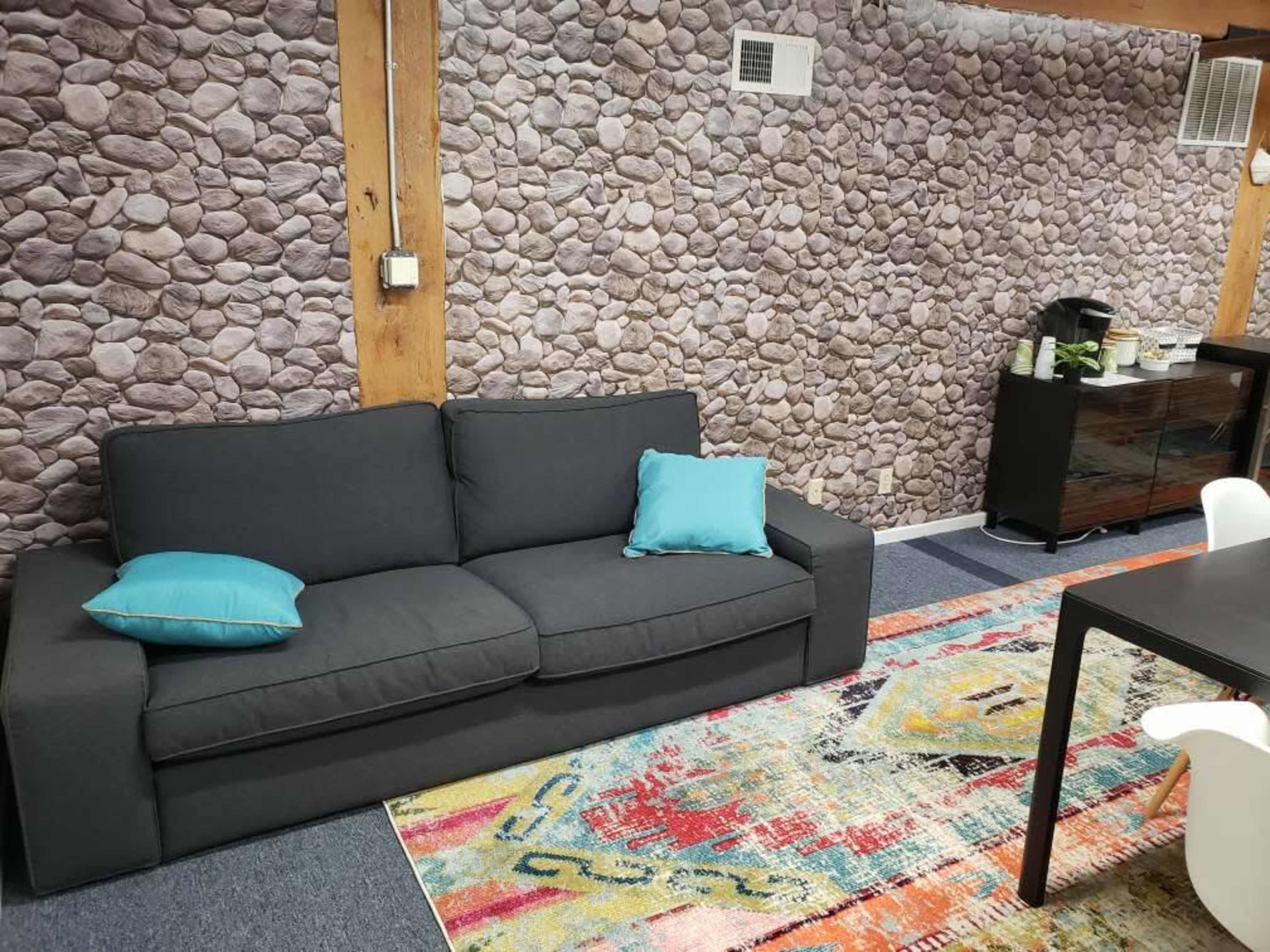 A living space with a gray sofa adorned with two turquoise cushions, a colorful patterned area rug, and a stone-patterned wall in the background.