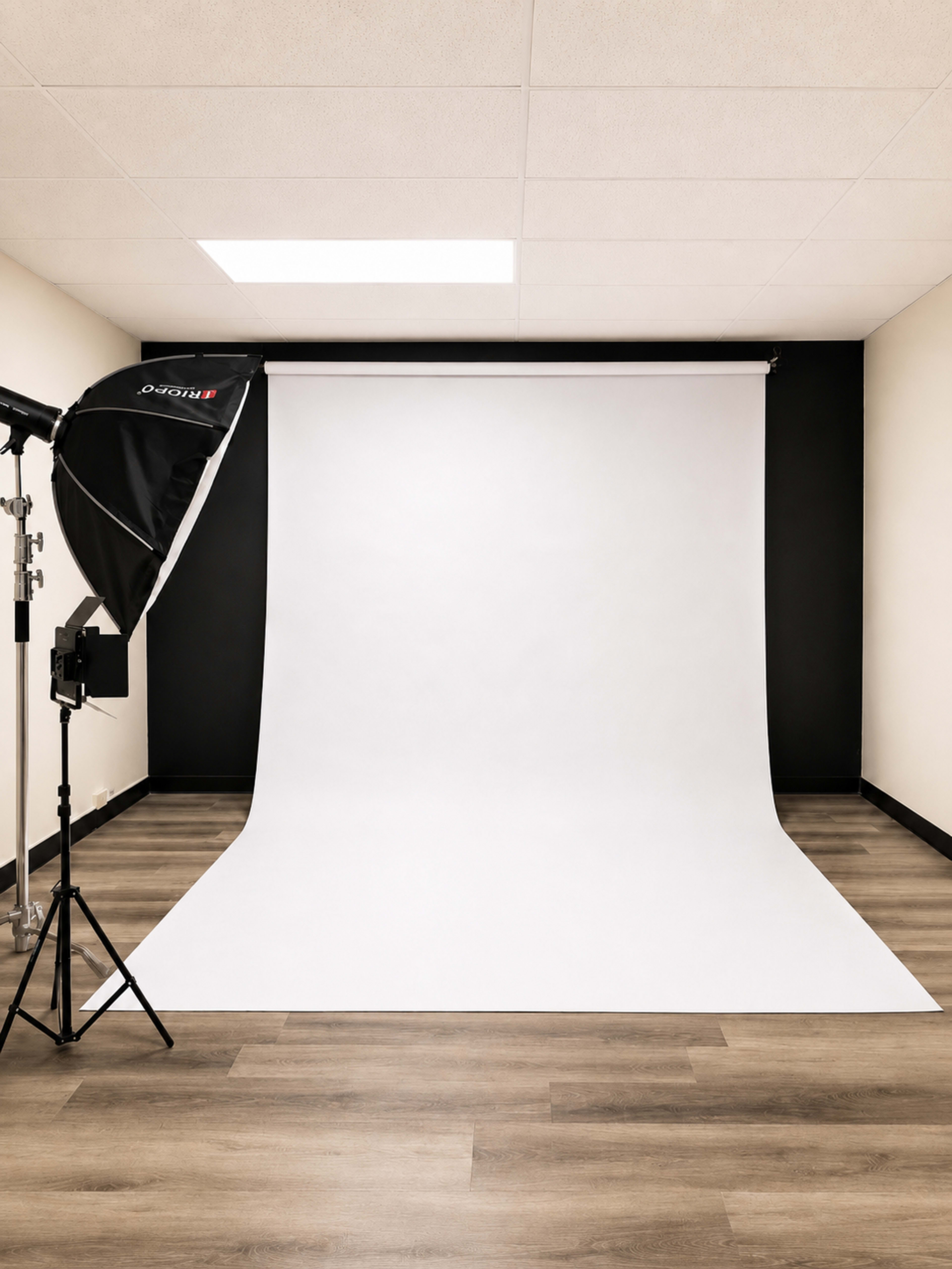 The image shows a photography studio with a large white backdrop, a softbox light, and wood flooring.