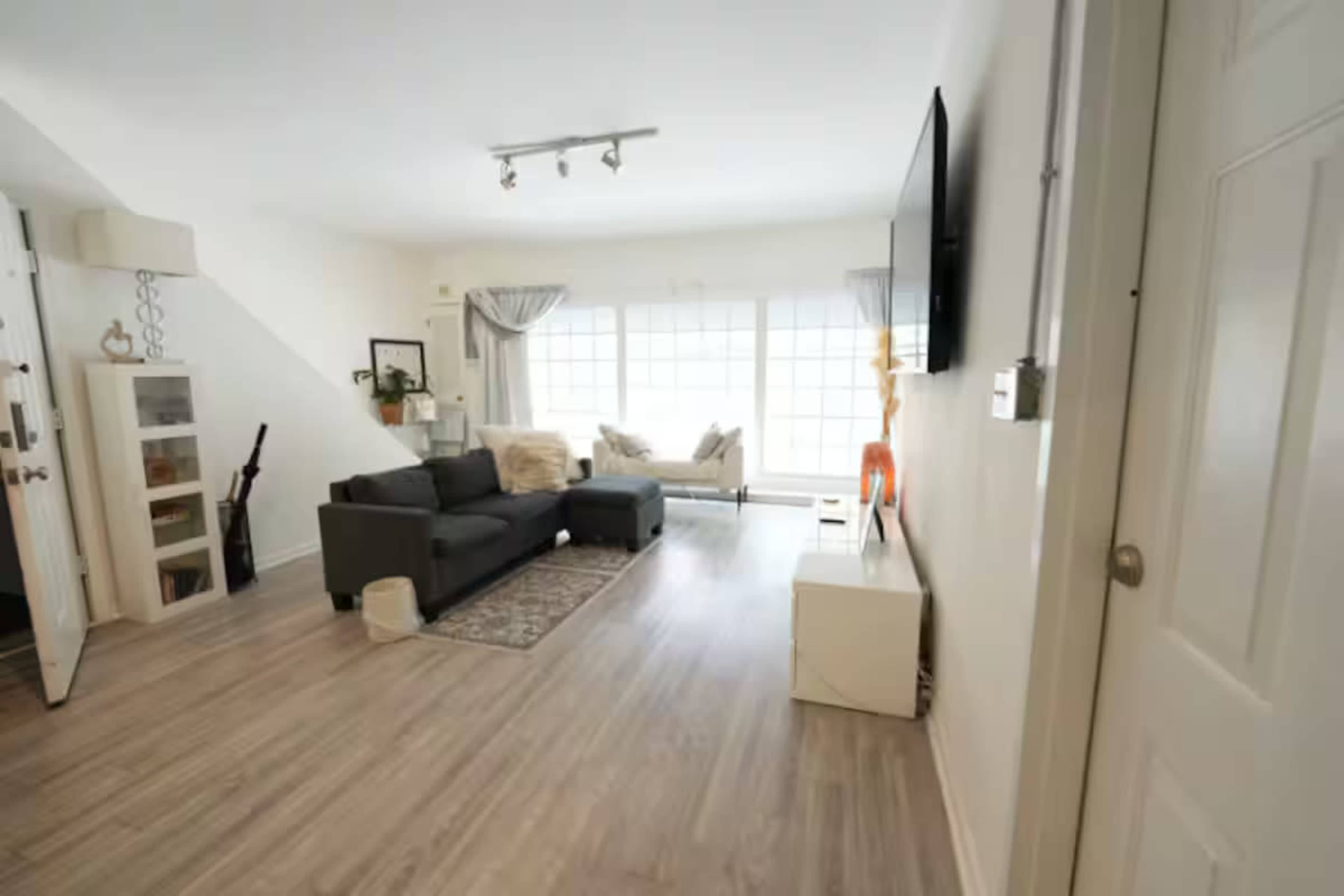 The image shows a modern living room with a gray sofa, a white television cabinet, and large windows allowing natural light to fill the space.