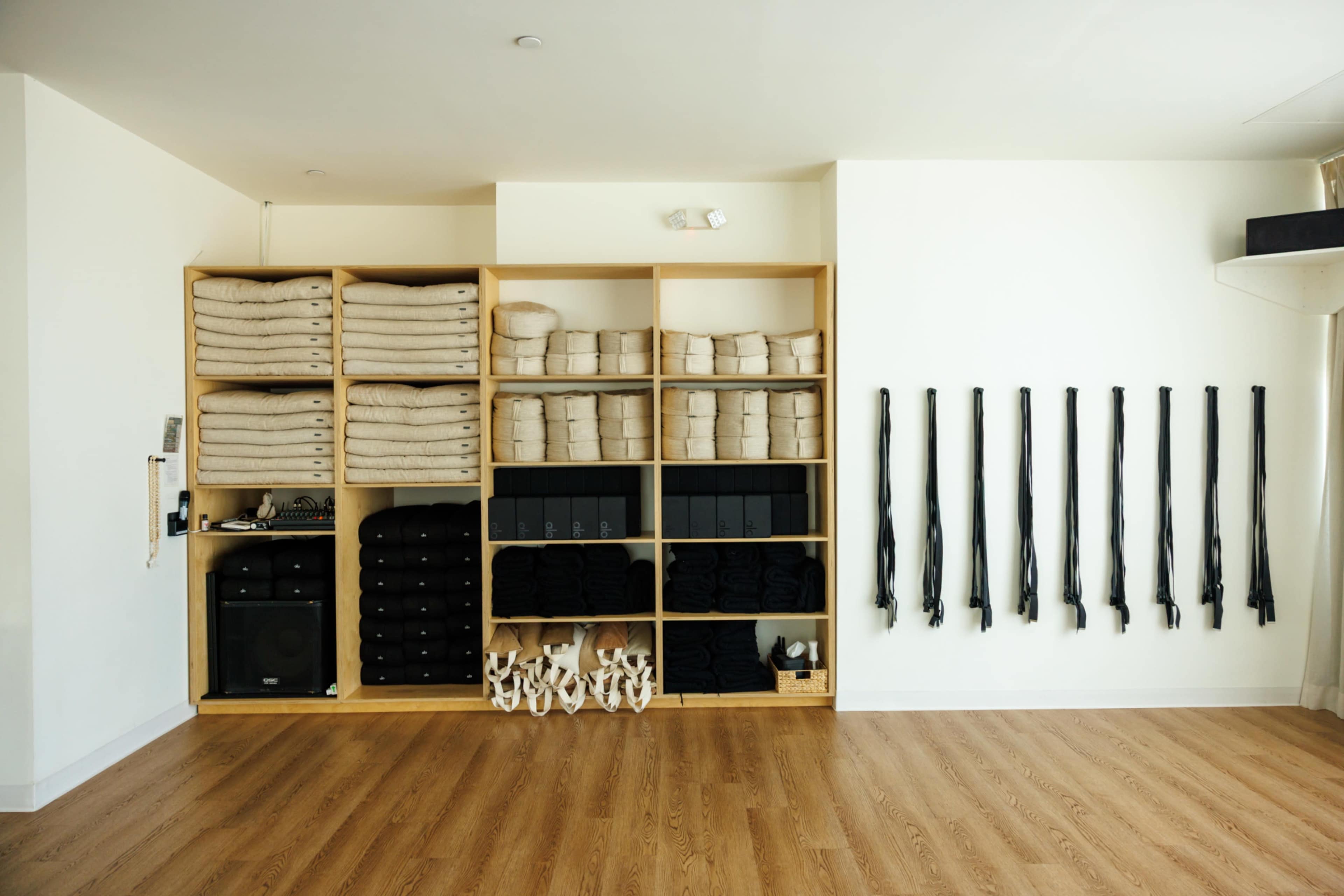 The image shows a neatly organized interior space featuring a wooden shelving unit filled with folded blankets and black yoga accessories, alongside rows of black straps hanging against a white wall.