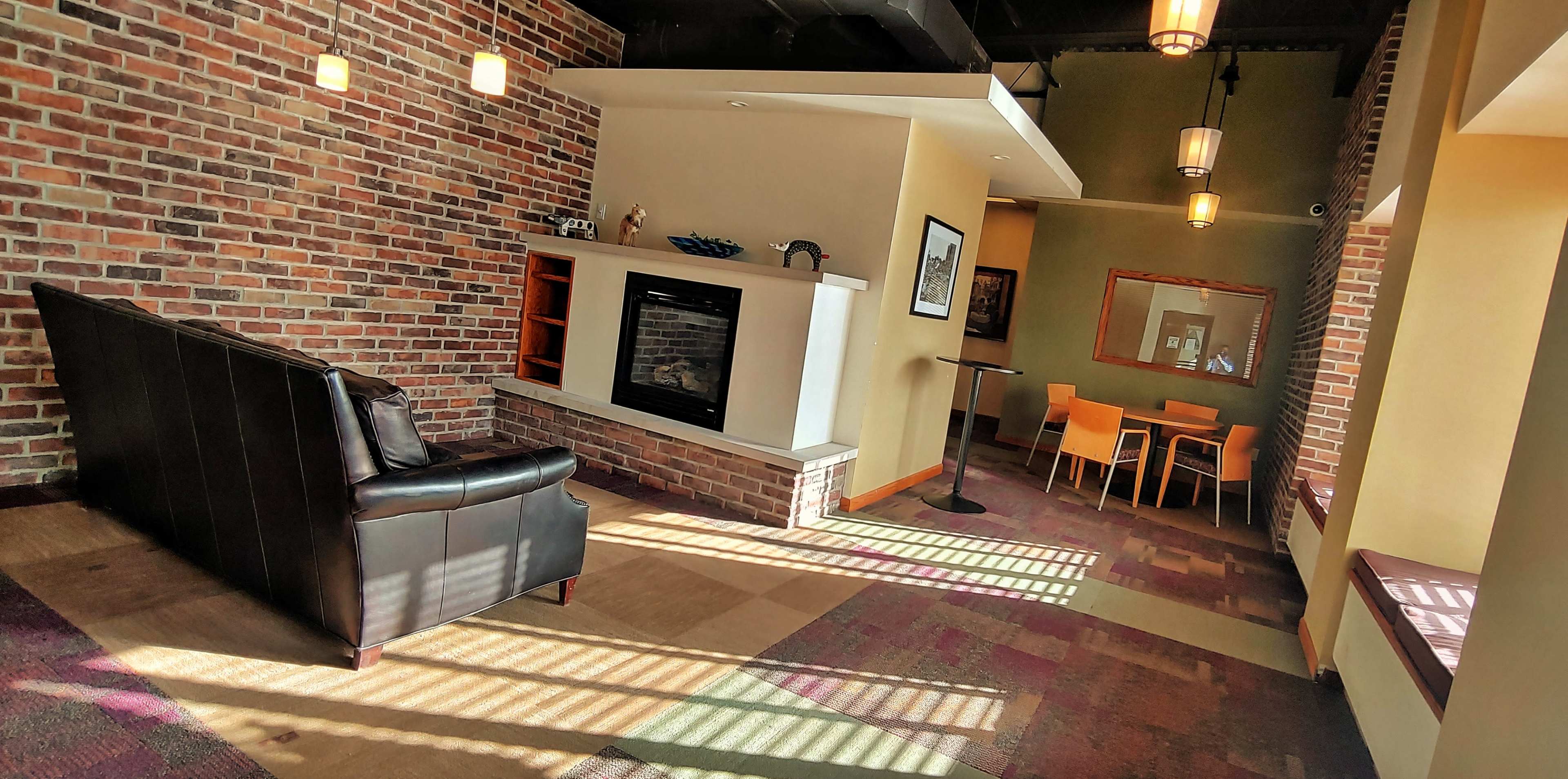 A cozy seating area with a black couch, a brick accent wall, a fireplace, and a dining table set against a backdrop of warm sunlight streaming through the windows.