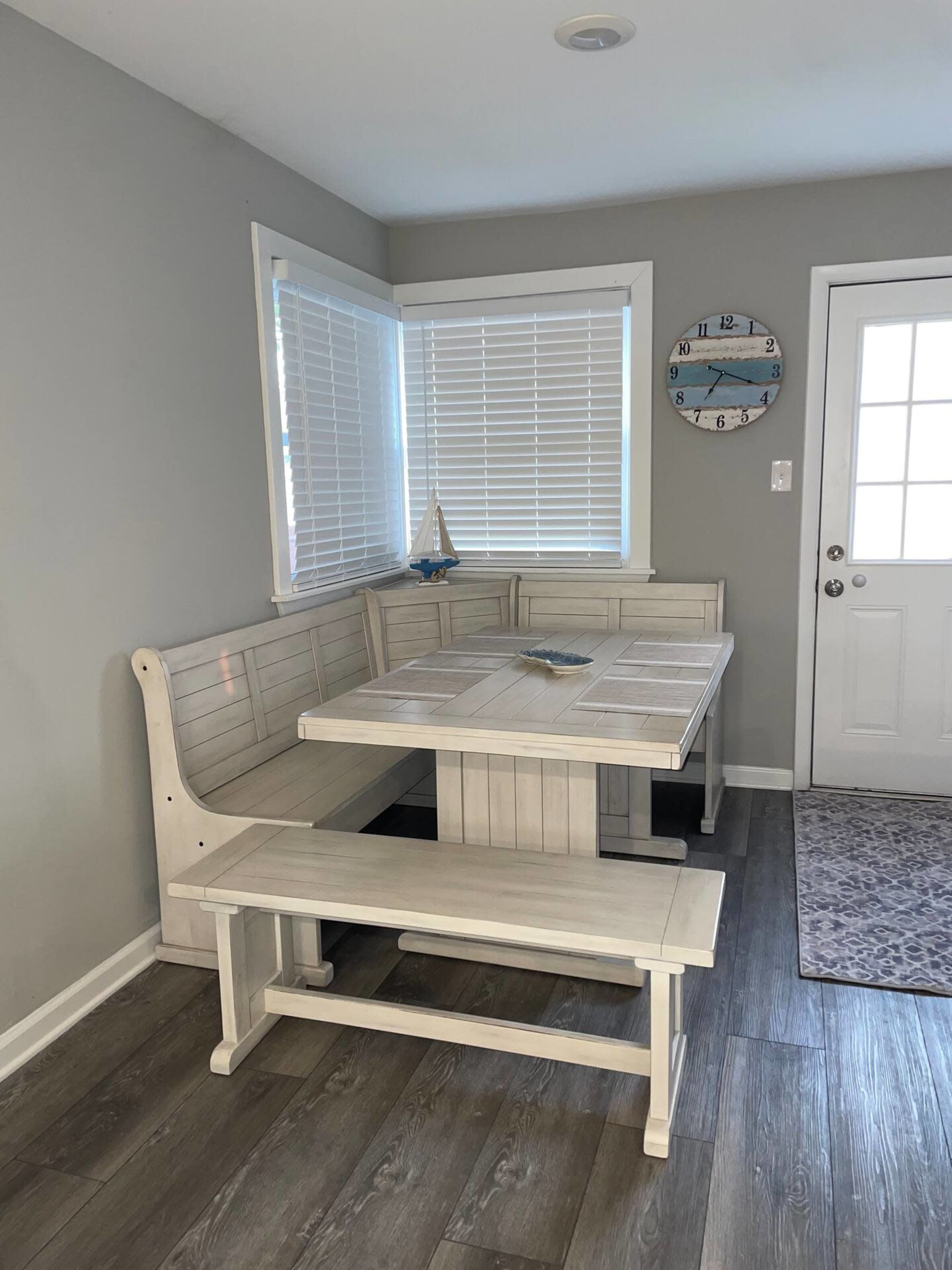 A light-colored wooden dining set with a table and benches is positioned in a corner near a door and windows with blinds.