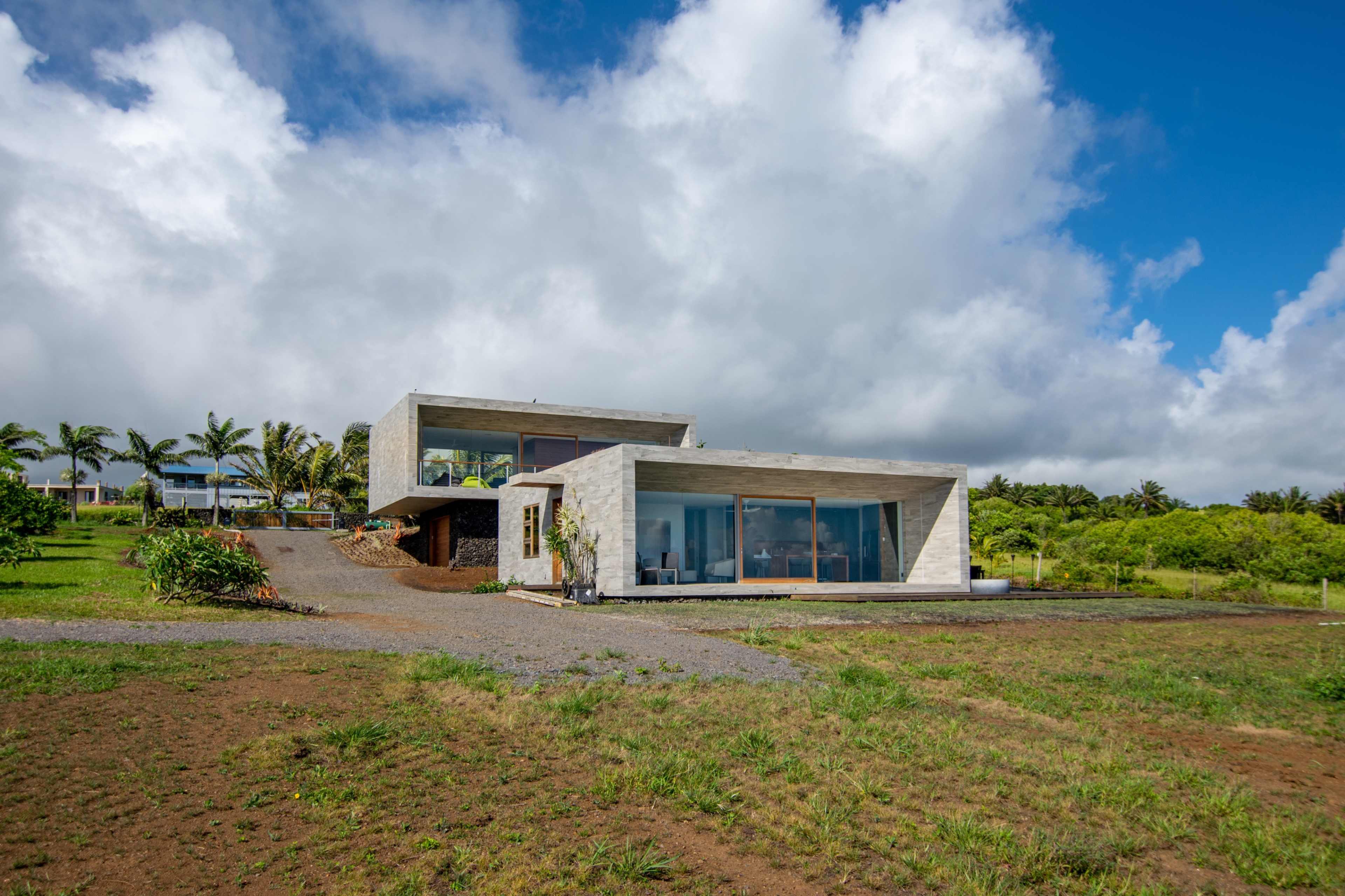 A modern concrete house with large windows is situated on a grassy landscape under a partly cloudy sky.