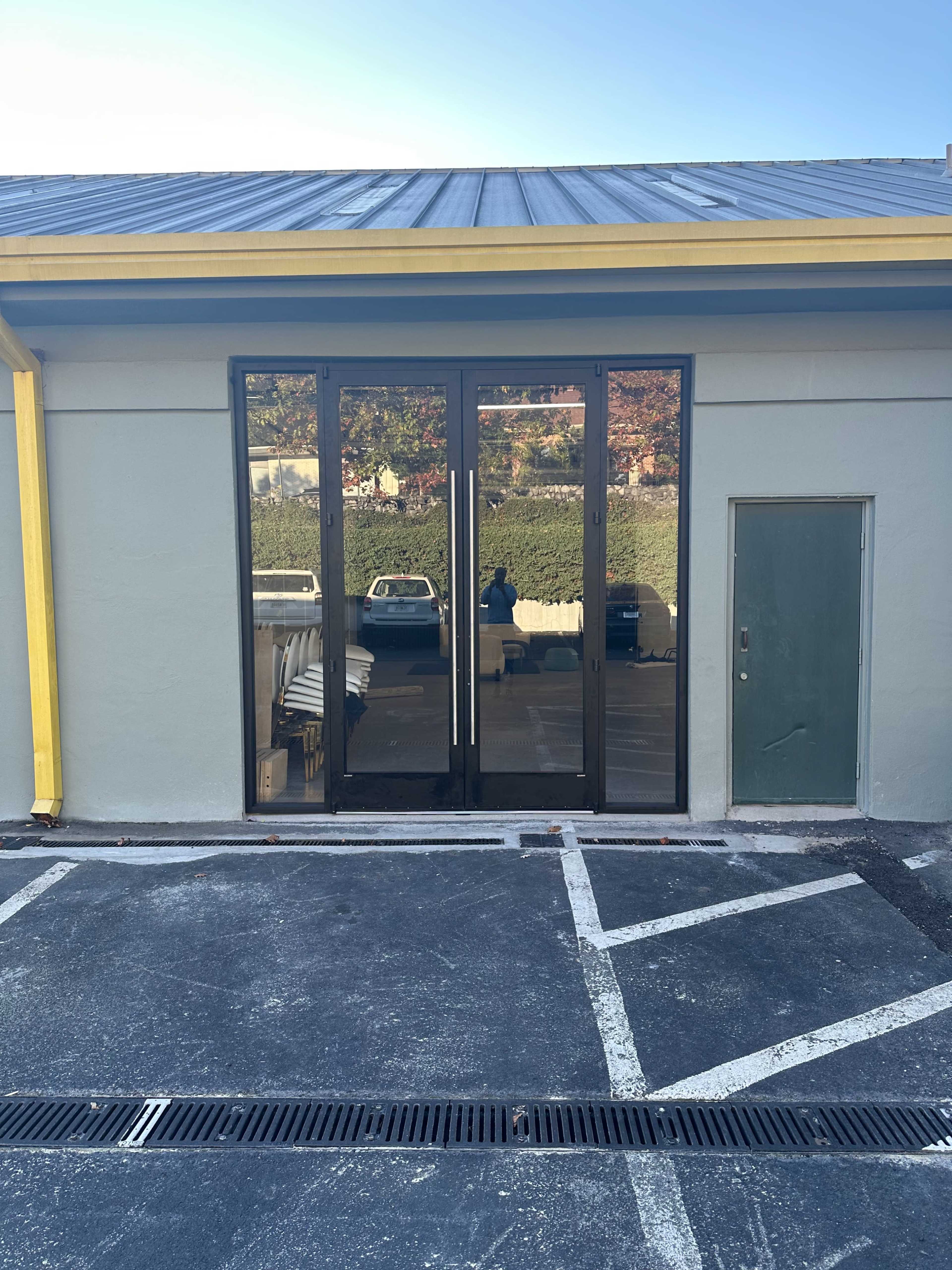 A building facade featuring large glass doors with a muted gray wall and a side door, all situated next to a parking lot.