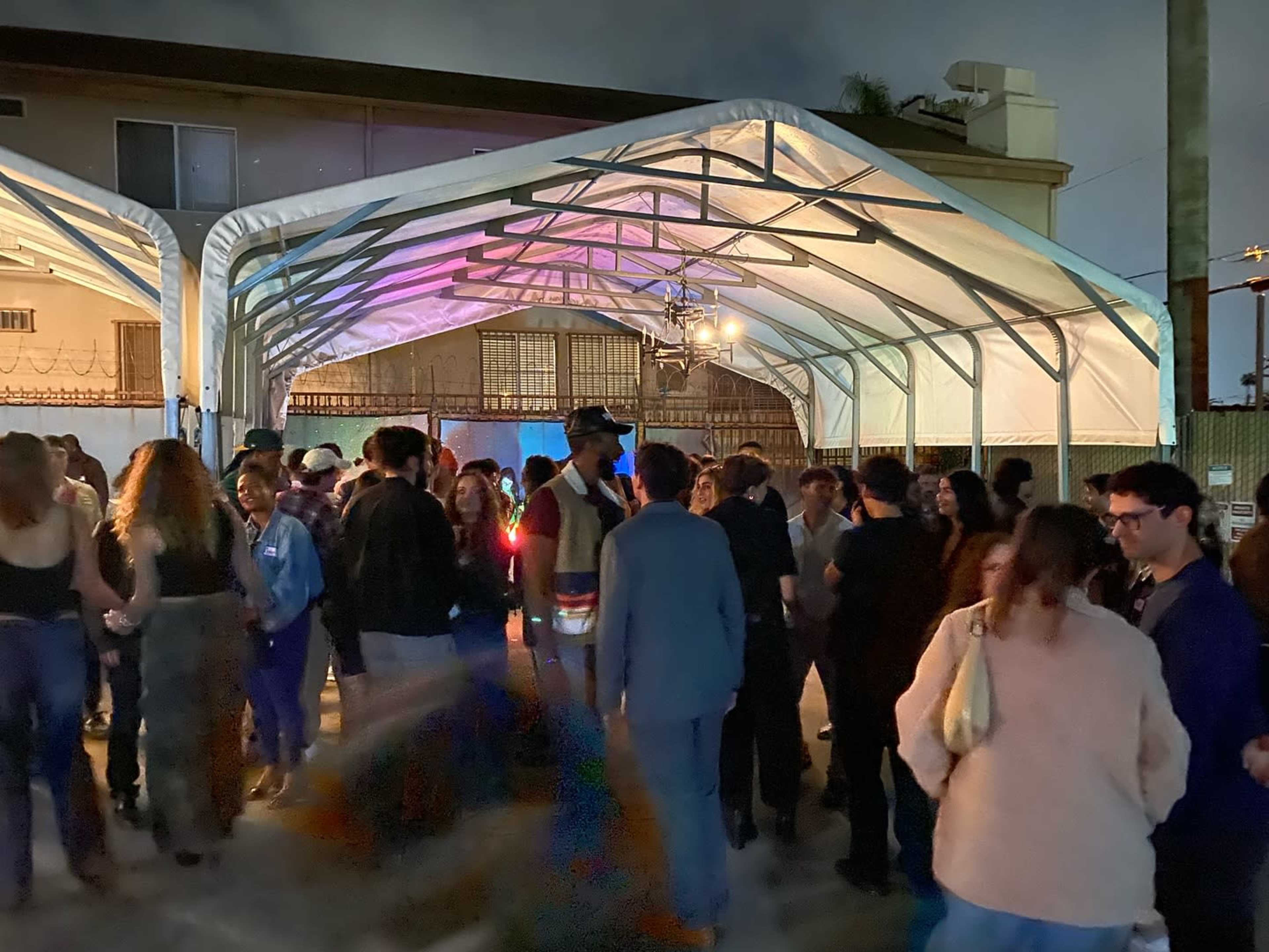 A large gathering of people is taking place under a tented structure at night, with colorful lights illuminating the scene.