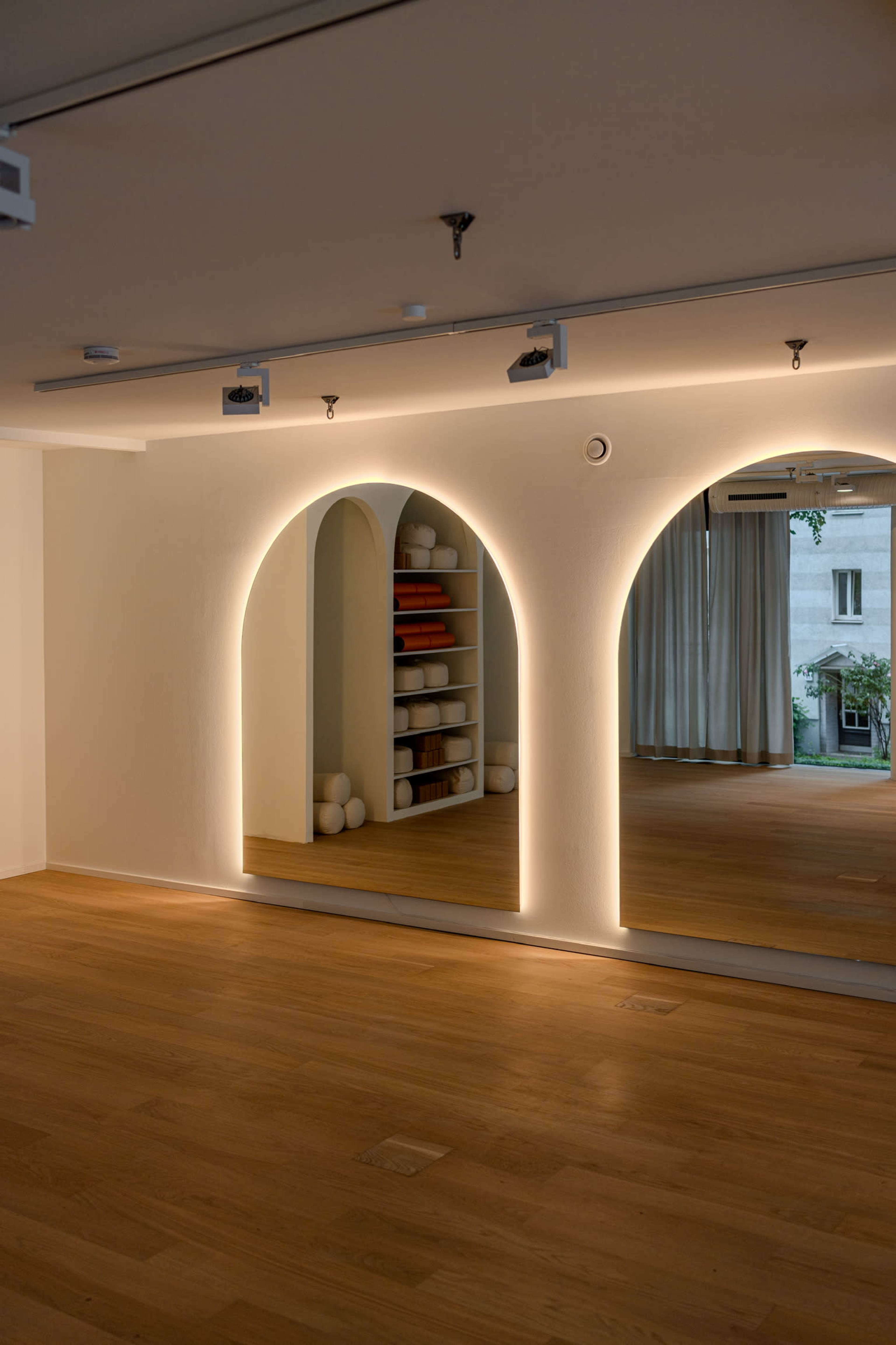 The image shows a minimalist room with wooden flooring, featuring arch-shaped mirrors illuminated by soft lighting and shelves in the background.