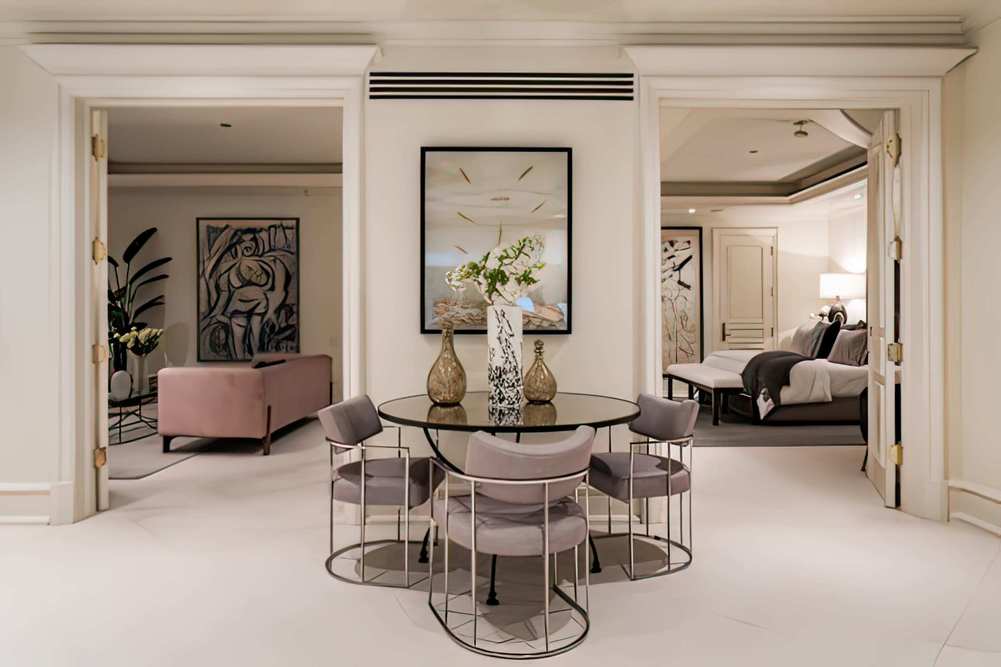 The image shows a modern dining area with a circular glass table surrounded by upholstered chairs, positioned between two doorways leading to a living space and a bedroom.