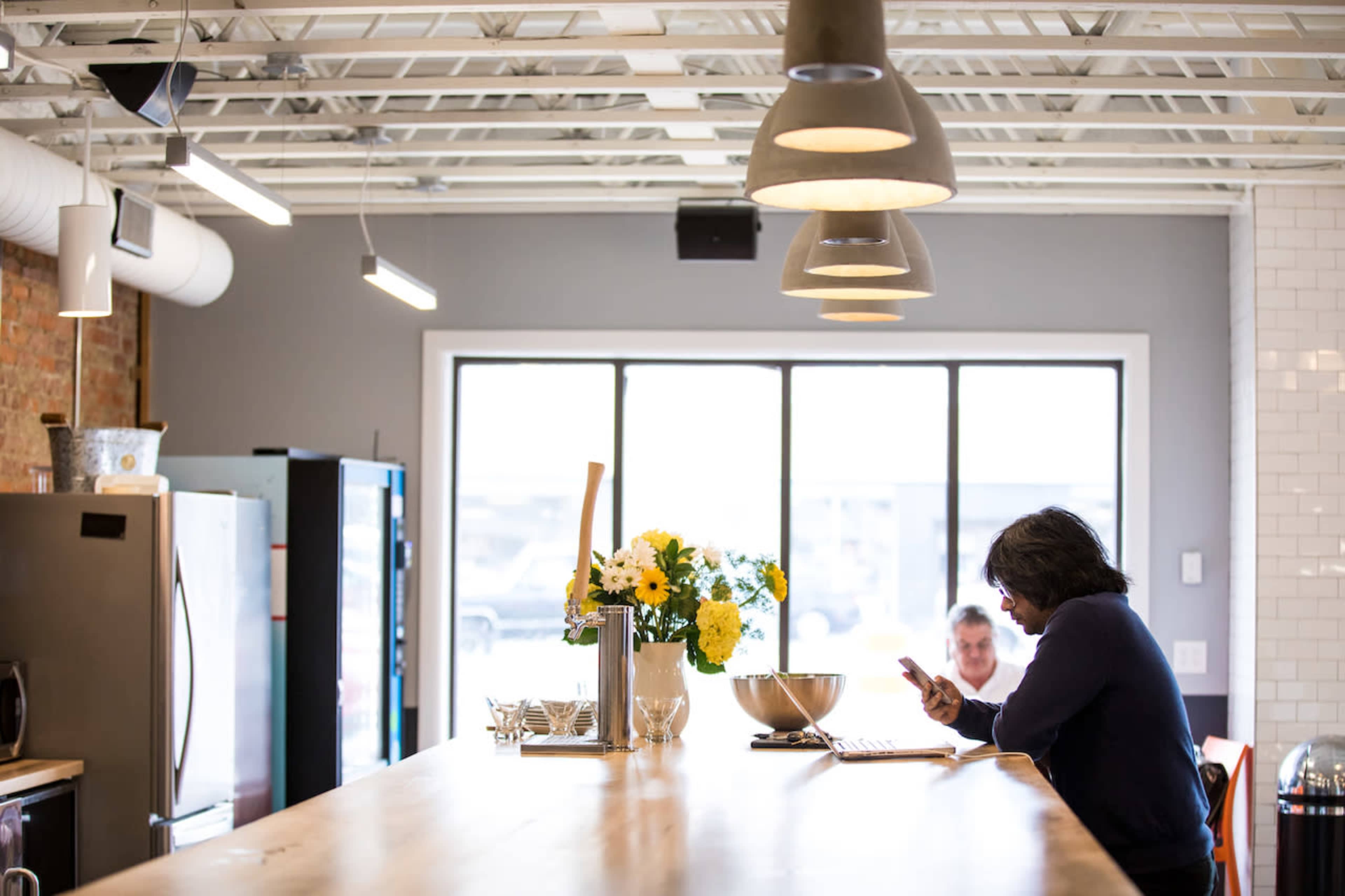 A person sits at a wooden table in a bright kitchen, looking at their phone while a vase of yellow flowers sits in the center.