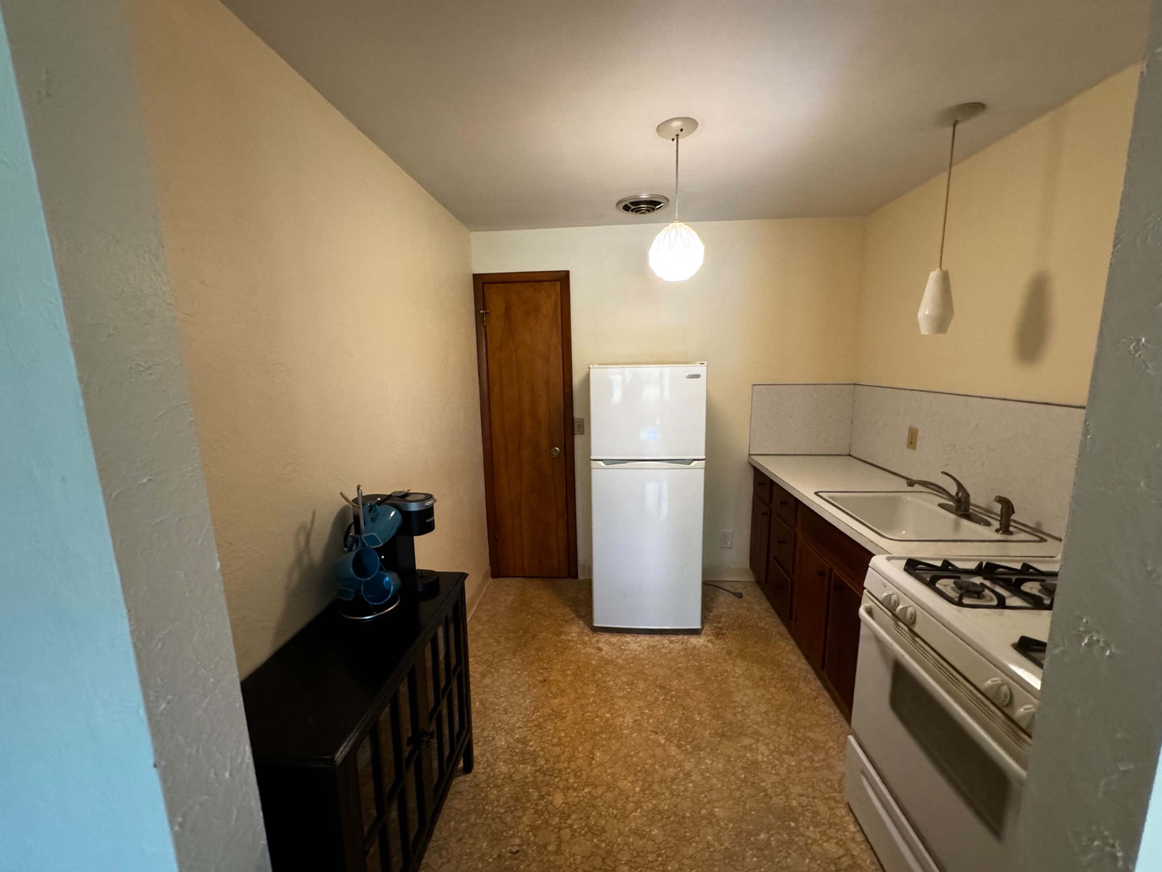 A small kitchen features a refrigerator, stove, and sink, with a doorway leading to another room and a pendant light hanging from the ceiling.