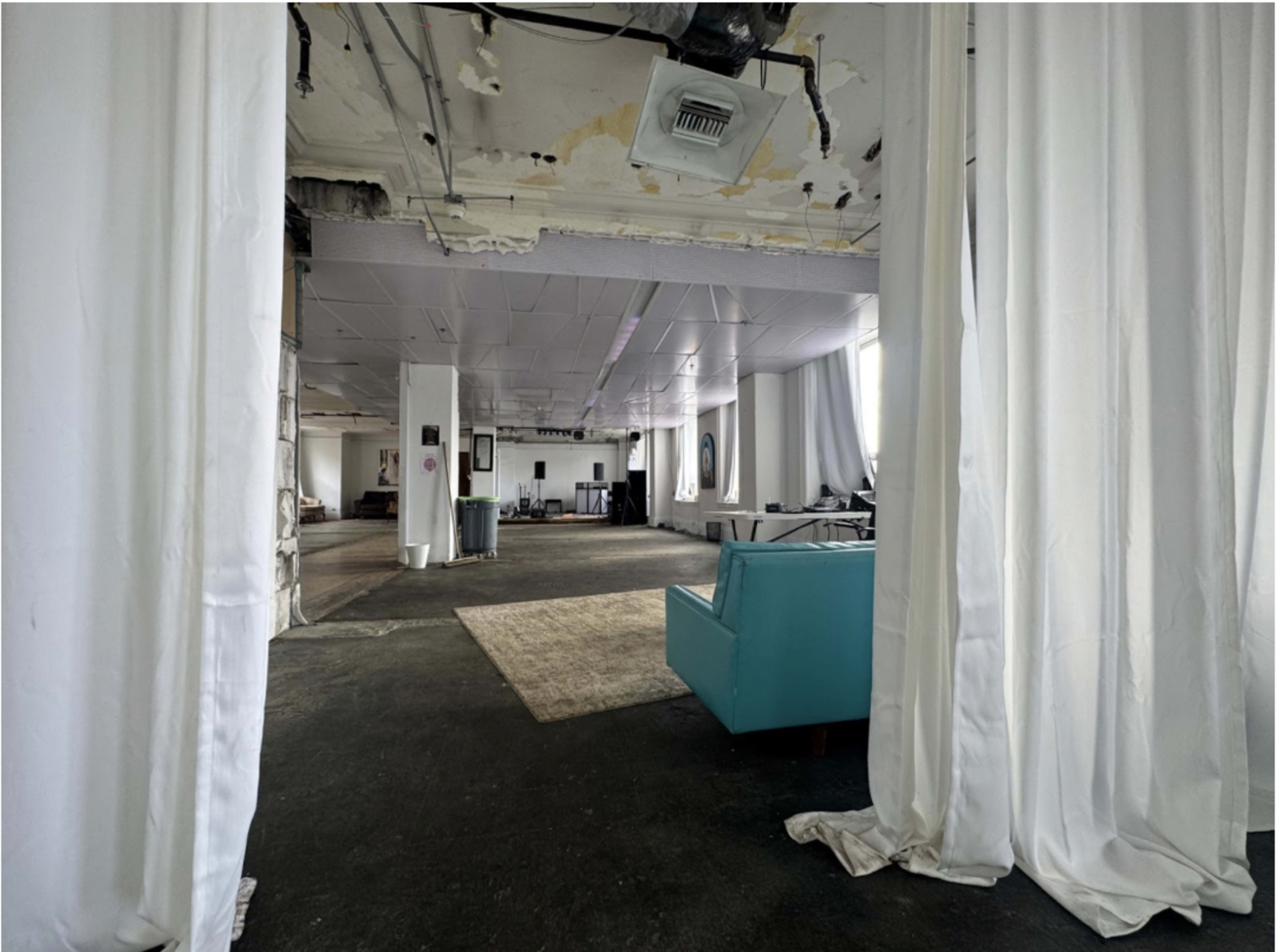 The image shows a spacious, partially furnished industrial interior with white drapes, a turquoise couch, and an open layout.