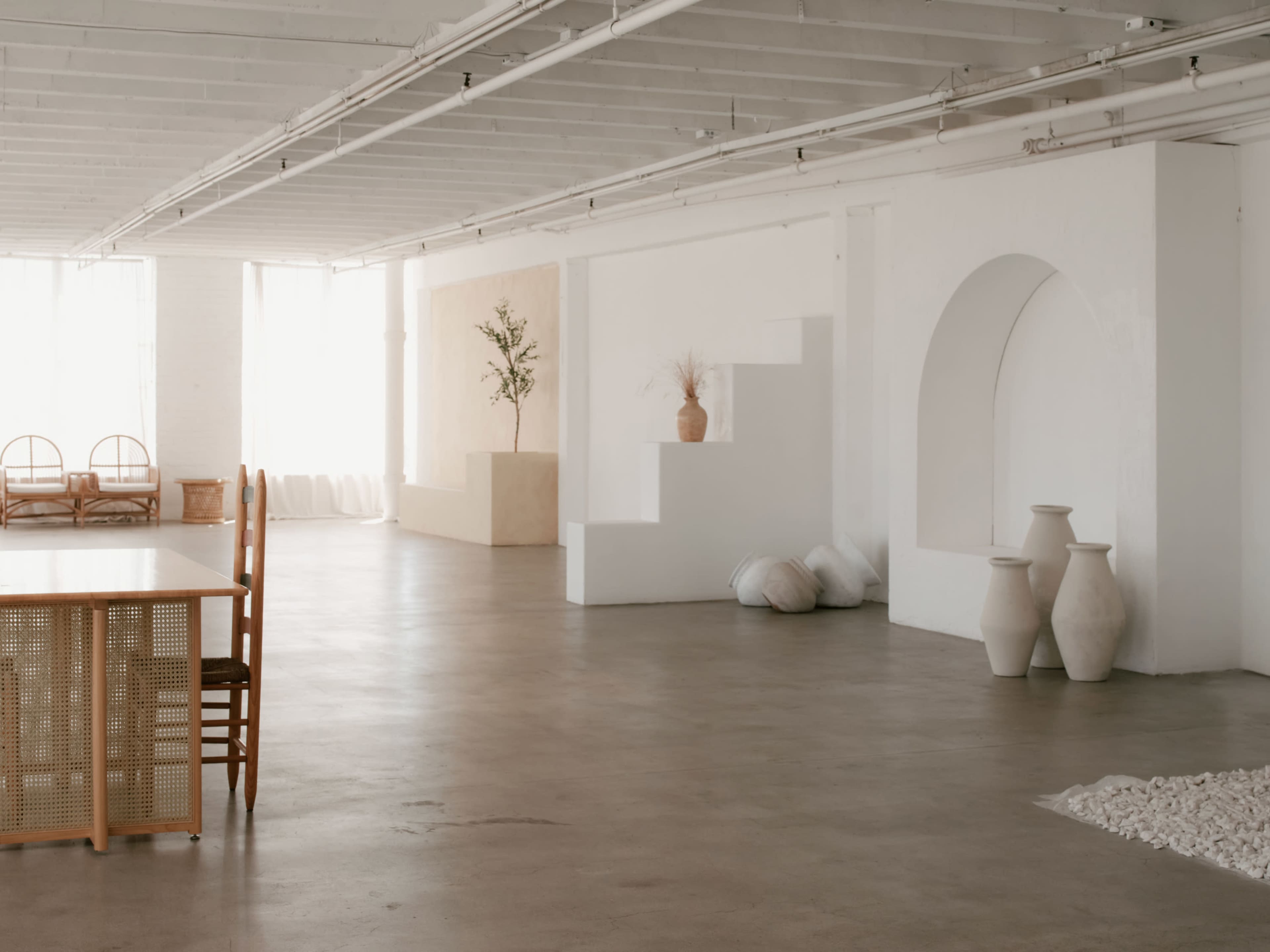 The image depicts a minimalist, well-lit interior space with white walls, a concrete floor, a dining table with chairs, and various decorative elements like plants and sculptural forms.