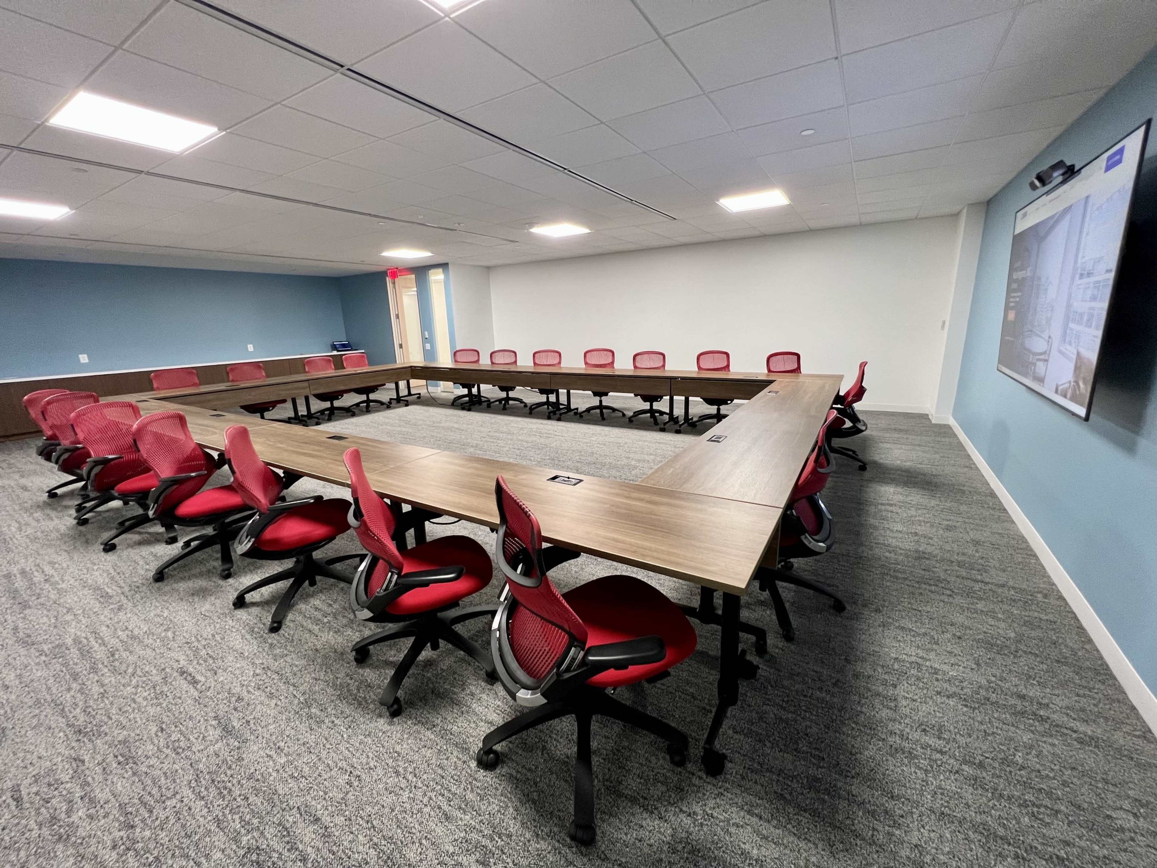 A large, rectangular conference table is arranged with red office chairs around it in a well-lit meeting room.
