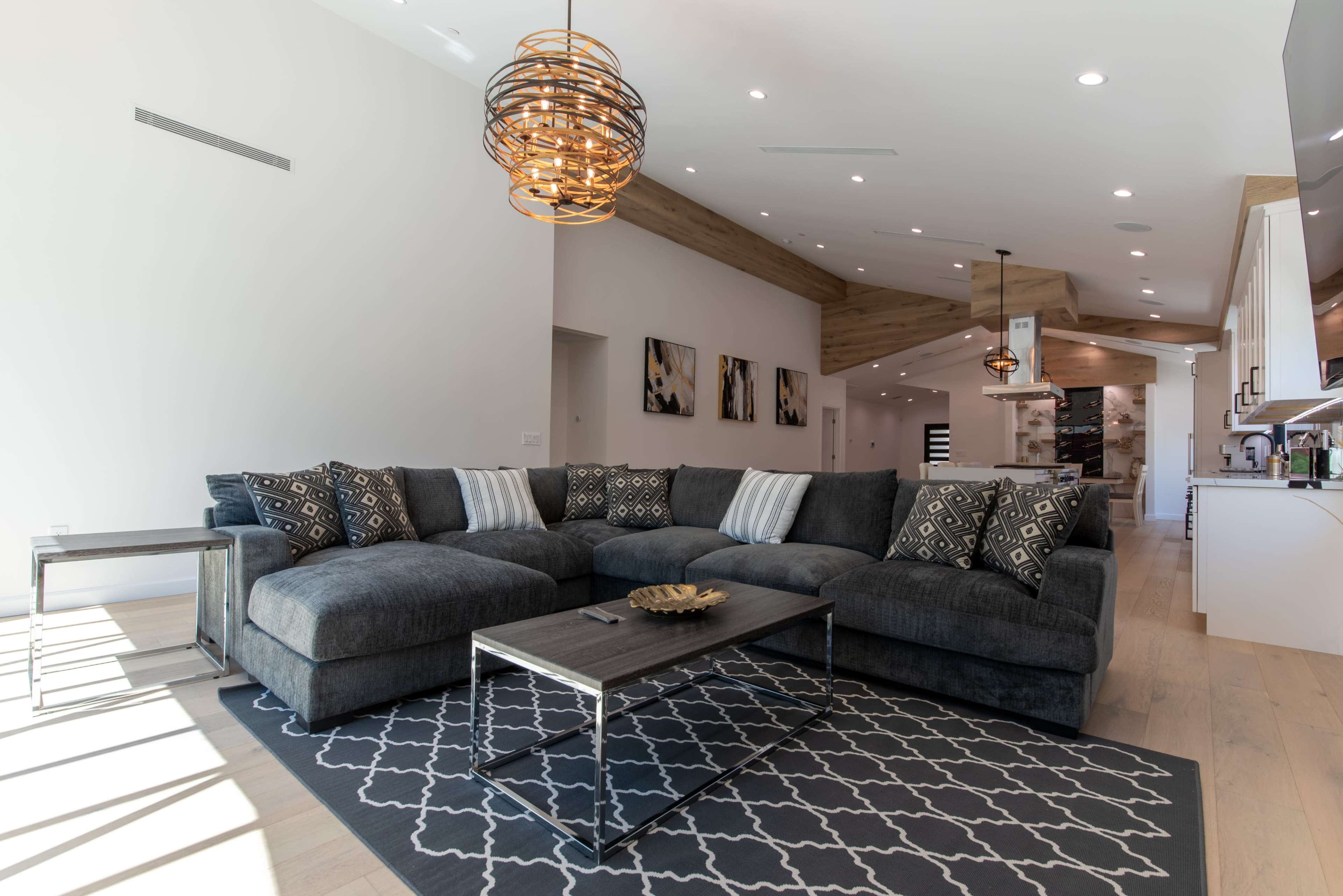 A spacious living room features a large sectional sofa, a low coffee table, and modern lighting fixtures, with a visible kitchen area in the background.